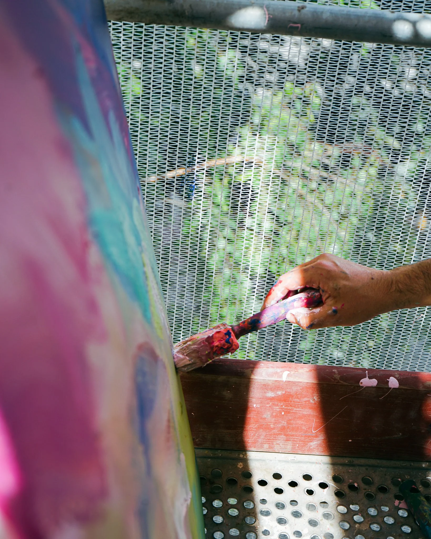 Nahaufnahme einer Hand, die mit bunten Farben auf einem Pinsel malt, vor einem gemalten Kunstwerk auf einer Leinwand im Freien.