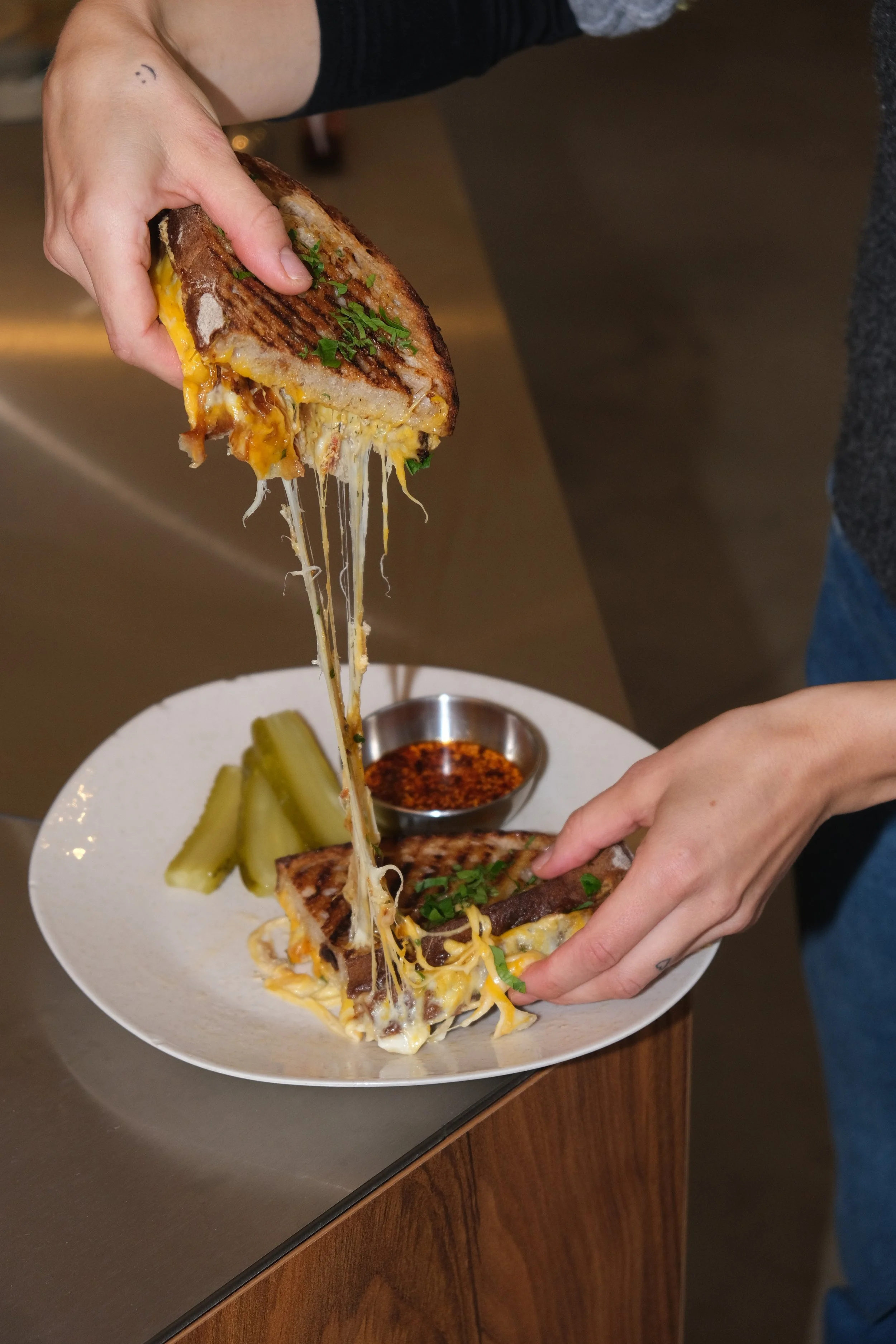 Nahaufnahme eines Sandwiches mit geschmolzenem Käse, das gerade halbiert wird. Auf einem Teller mit Gewürzgurken und einer kleinen Schüssel mit roter Soße. Hände, die das Sandwich halten.