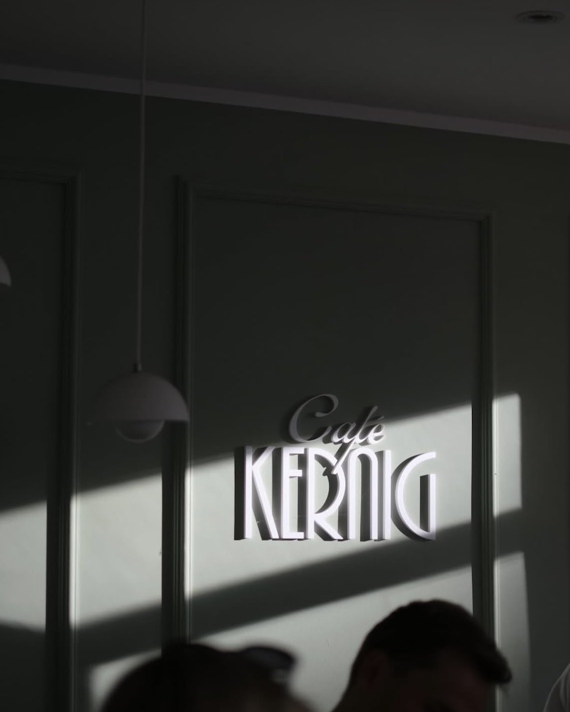 Innenraum eines Cafés mit einem beleuchteten Schriftzug 'Cafe KERMTIC' an der Wand, Schatten und Oberlicht