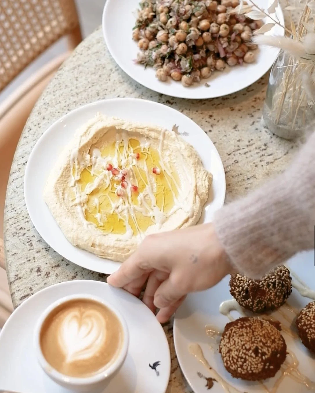 Verschiedene süße Speisen auf einem gedeckten Tisch, darunter Hummus mit Olivenöl und Granatapfelkernen, eine Schale mit Kichererbsensalat, ein Cappuccino mit Herz-Muster, und drei Schokoladenküchlein mit Sesam bestreut.