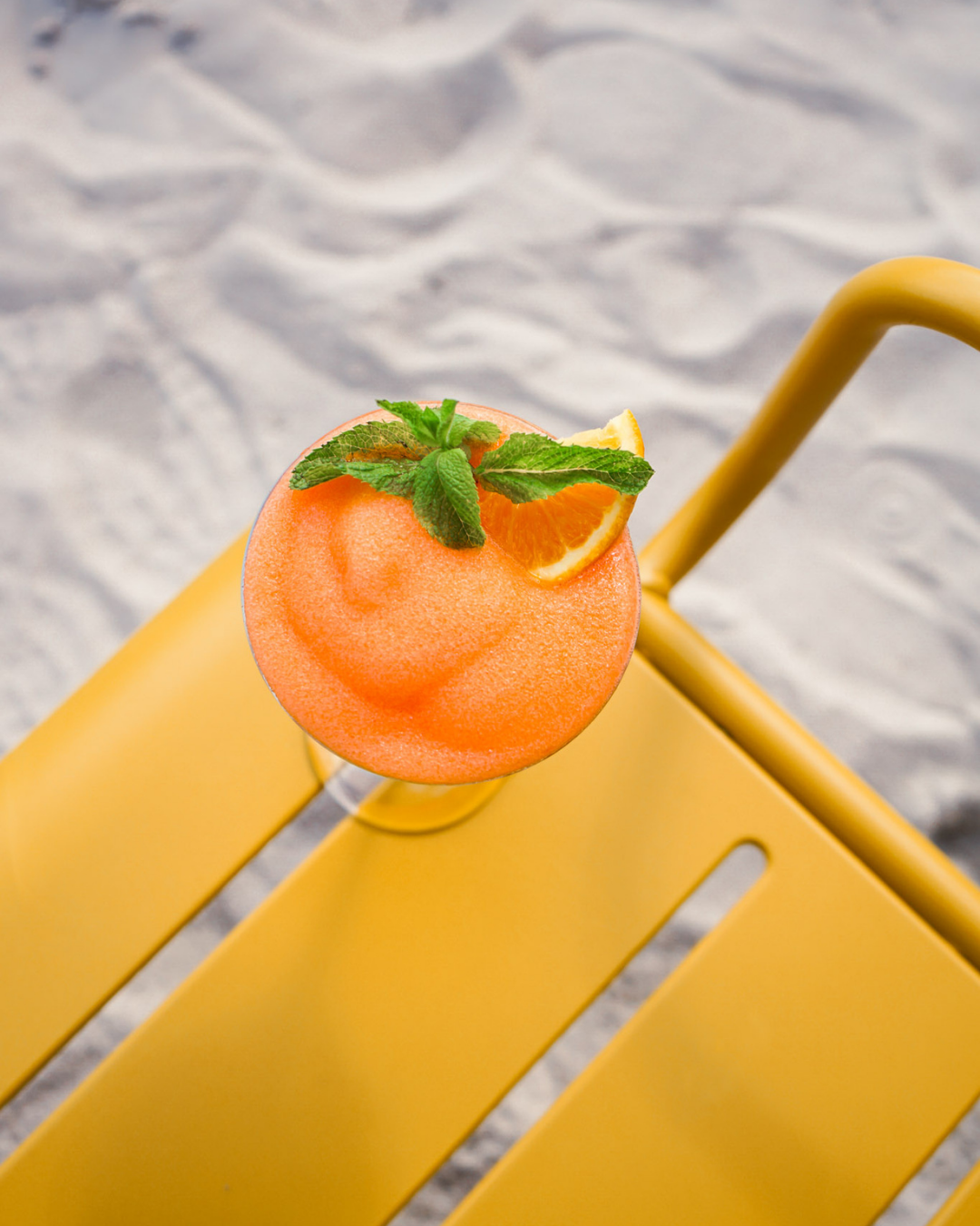 Ein orangefarbener Früchte-Cocktail mit Minzblättern, Orangenscheibe und Strohhalm auf einem gelben Tisch im Sand
