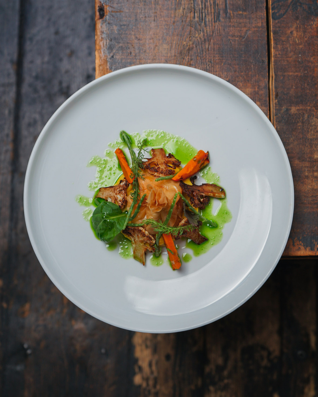 Ein weißer Teller mit einem Gourmet-Gericht aus gebratenen Gemüsesorten, grüner Soße, Gemüse und einer dünnen Teigscheibe, auf einem Holztablett