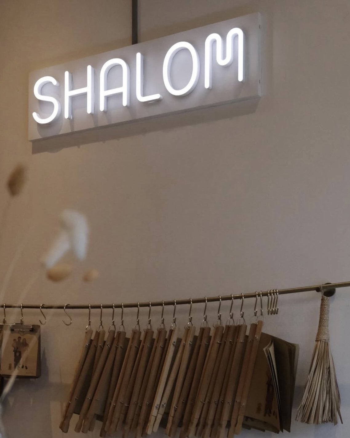 Neon-Schriftzug mit der Aufschrift 'Shalom' an einer Wand, darunter eine Metallstange mit hängenden Holzstäbchen, neben einer Buchseite und einem Besen.