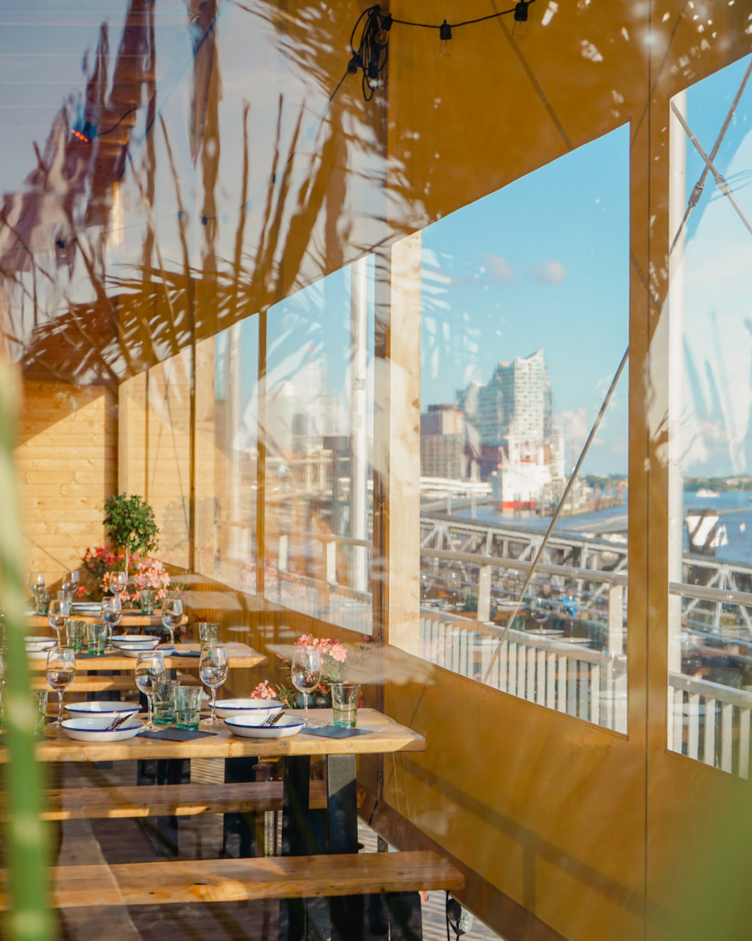 Blick durch eine Fensterwand auf einen Speisetisch mit Weingläsern und Blumen, mit Stadt im Hintergrund unter blauem Himmel.