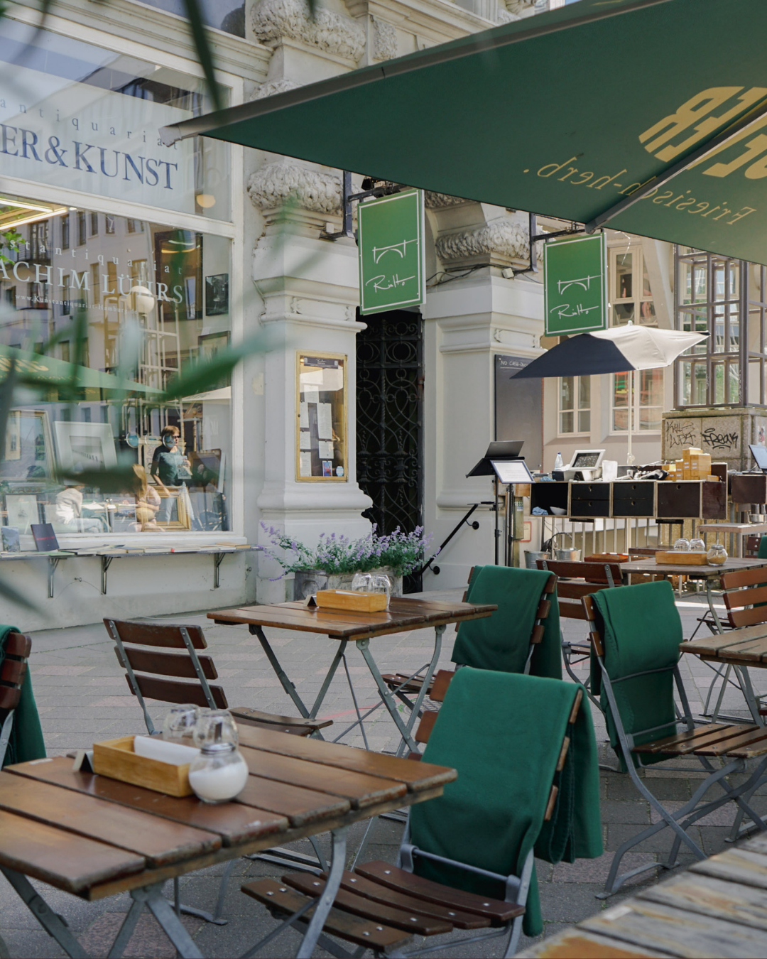Leerer Außenbereich eines Cafés oder Restaurants mit grünen Stühlen und Holztischen, Sonnenschirmen, und einem Geschäft im Hintergrund.