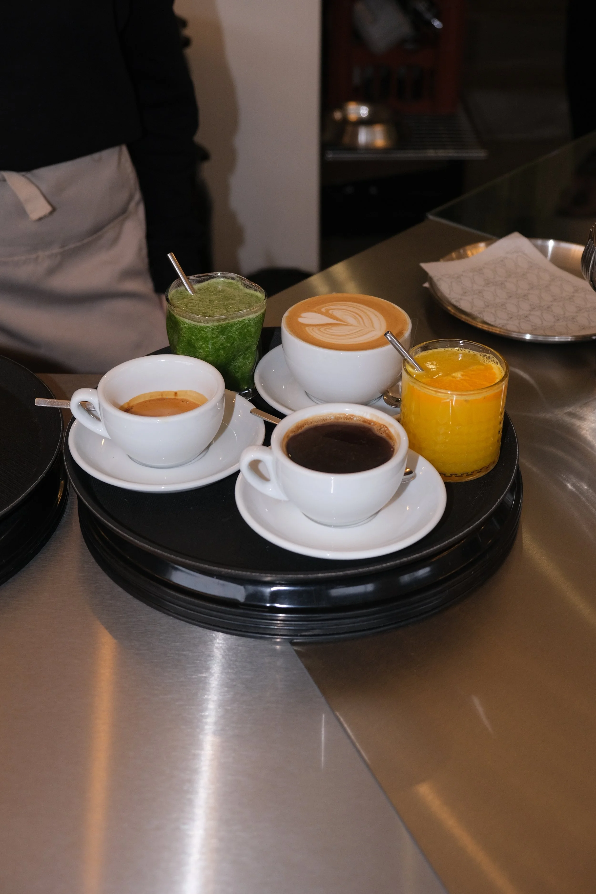 Verschiedene Kaffee- und Getränkearten auf einem schwarzen Tablett, darunter Cappuccino, Espresso, grüner Saft und Orangensaft, auf einer Metallarbeitsfläche in einer Küche oder Café.