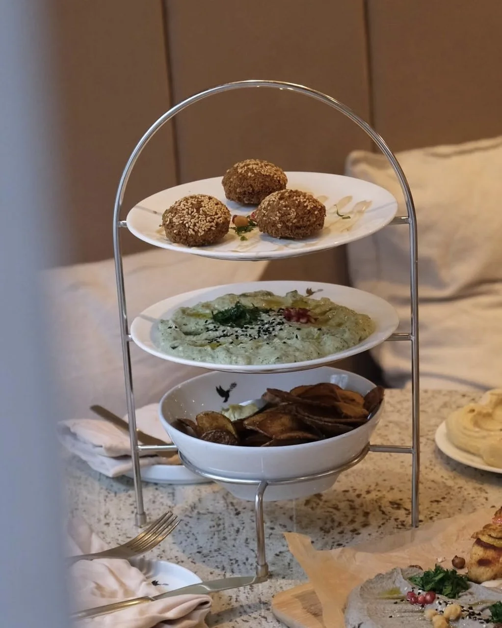 Dreistufiges Teeservice mit verschiedenen Speisen: oben drei gefüllte Kroketten, in der Mitte ein Teller mit grüner Sauce, unten eine Schale mit gebratenen Auberginen, auf einem Tisch mit Besteck und weiteren Speisen im Hintergrund.