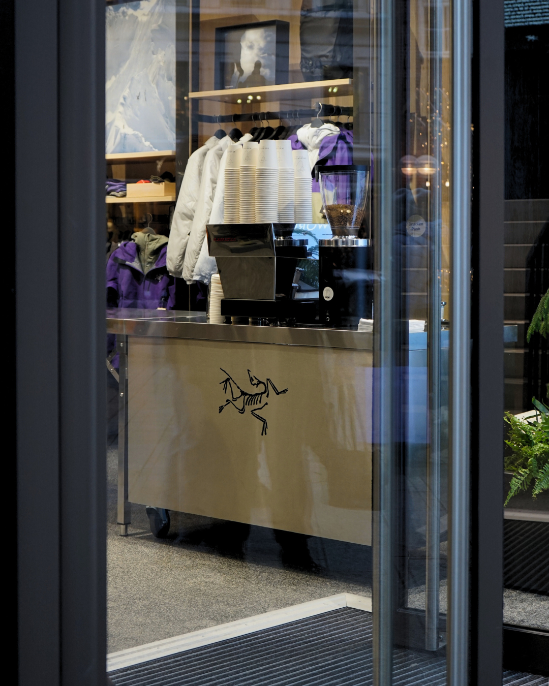 Innenansicht eines Kaffeestands in einem Geschäft mit einem Espressoautomaten, Kaffeebohnen im Kühler, Stapeln von Pappbechern und Kleidung im Hintergrund.