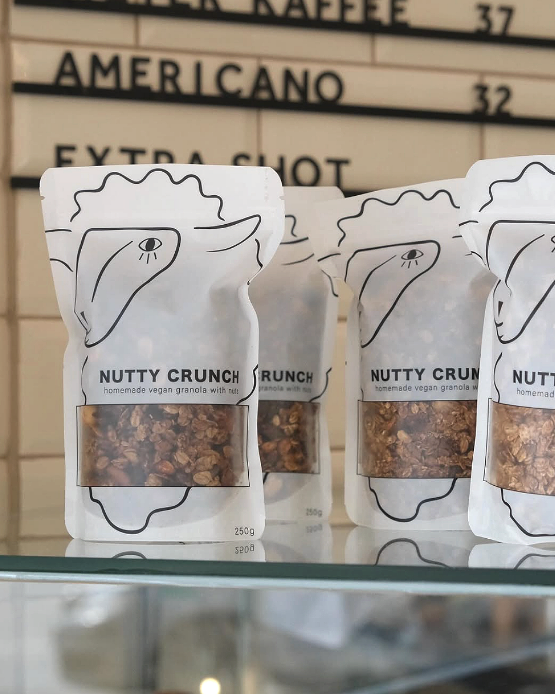 Vier weiße Verpackungen mit schwarzer Illustration eines Gesichts und offenem Mund, die Nutty Crunch veganes Granola mit Nüssen enthalten, stehen auf einer Glasregalfläche vor einem Menüboard.
