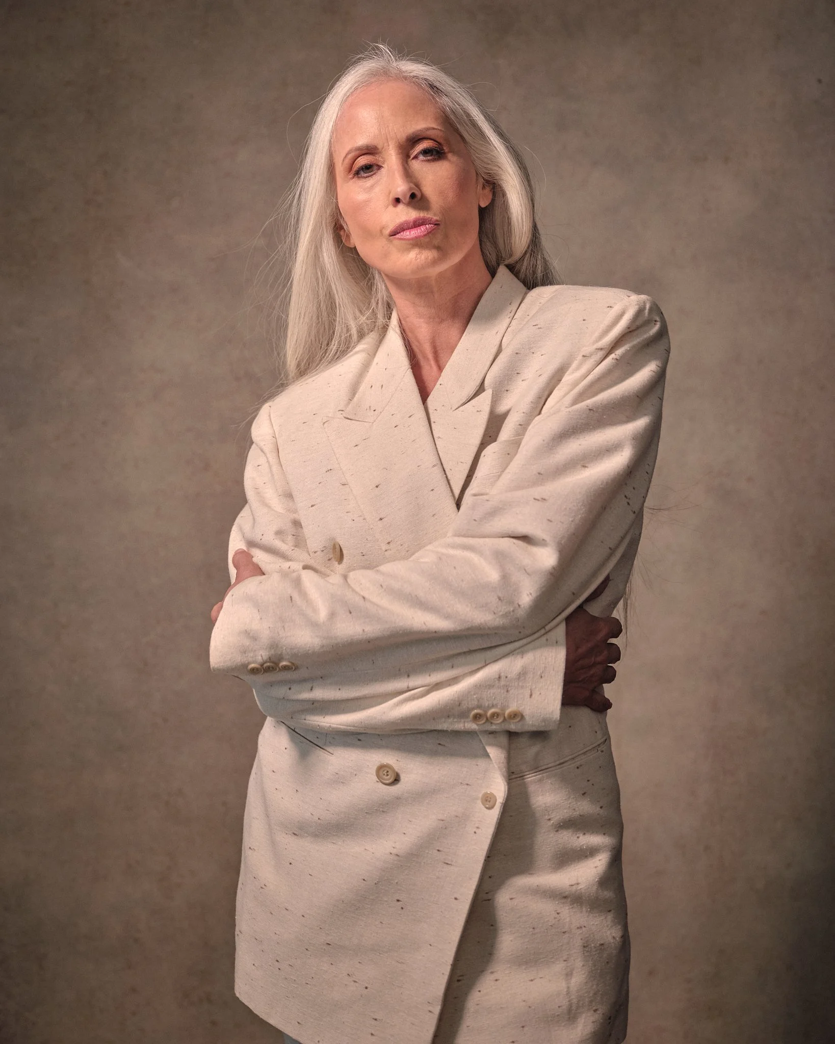 An elderly woman with long gray hair wearing a cream-colored blazer with arms crossed, standing against a plain background.