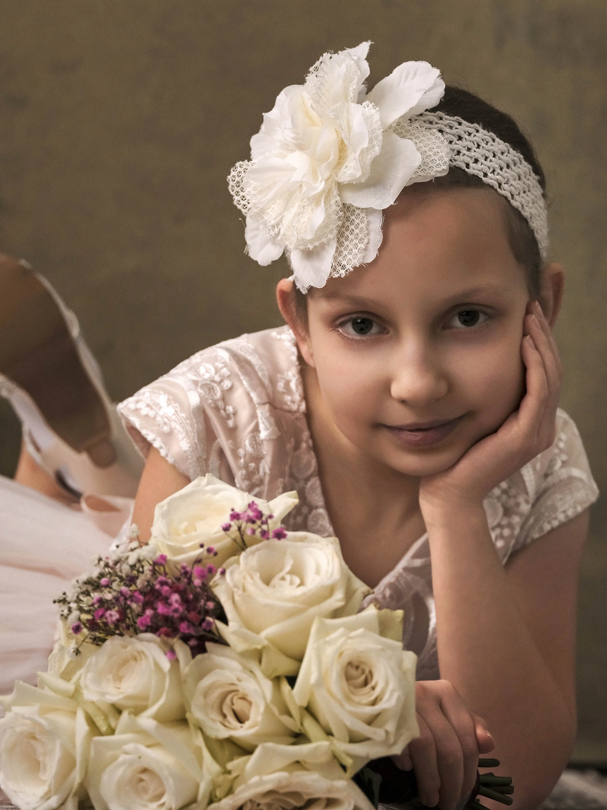A young girl with a white flower headband laying on her side, resting her face on her hand, near a bouquet of white roses and small pink flowers.