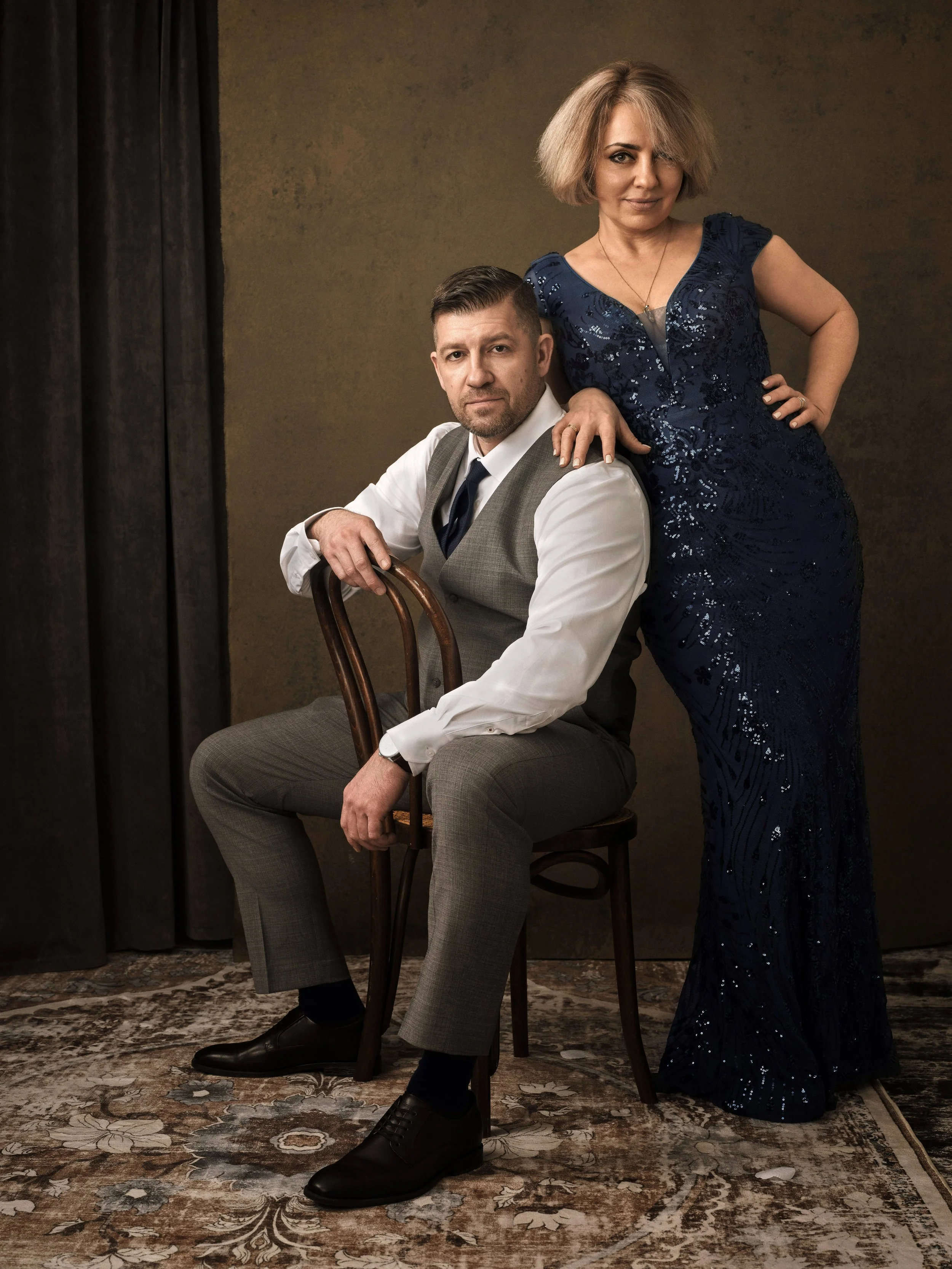 A man in a gray suit with a vest and tie sitting on a wooden chair, and a woman in a dark blue sequin gown standing behind him with her hand on his shoulder, posing for a formal portrait in front of a textured brown backdrop.