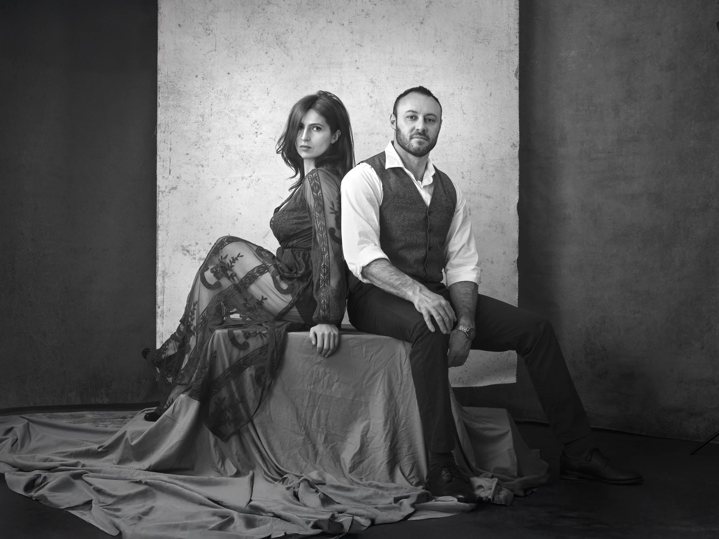 A black-and-white photograph of a man and woman sitting on a draped platform against a textured wall. The woman is wearing a sheer, embroidered dress and has shoulder-length hair, while the man is dressed in a vest over a button-down shirt and wearing a watch.
