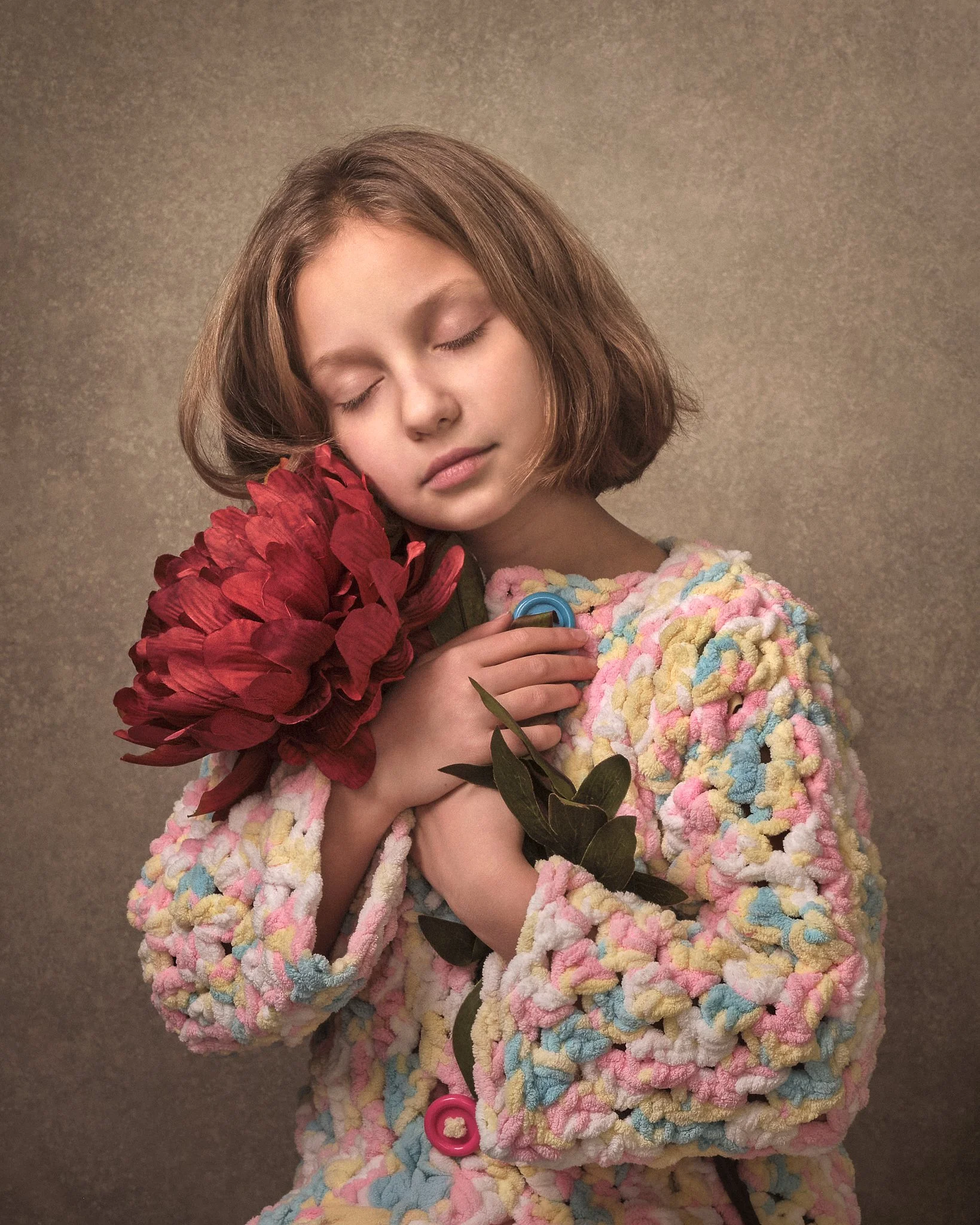 A young girl with short brown hair, closed eyes, and a peaceful expression, holding a large red flower close to her face, wearing a colorful textured coat, against a neutral background.