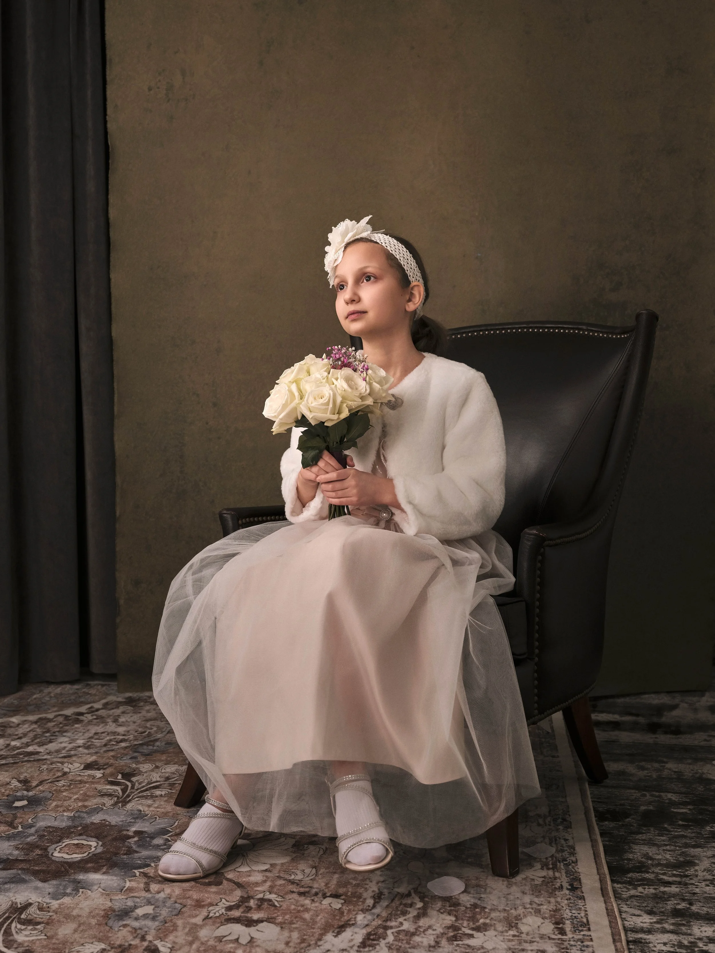 A young girl dressed in formal attire with a white dress, cardigan, and floral headband sitting in a black armchair, holding a bouquet of white roses, looking thoughtful, indoors with a brown wall and a patterned carpet.