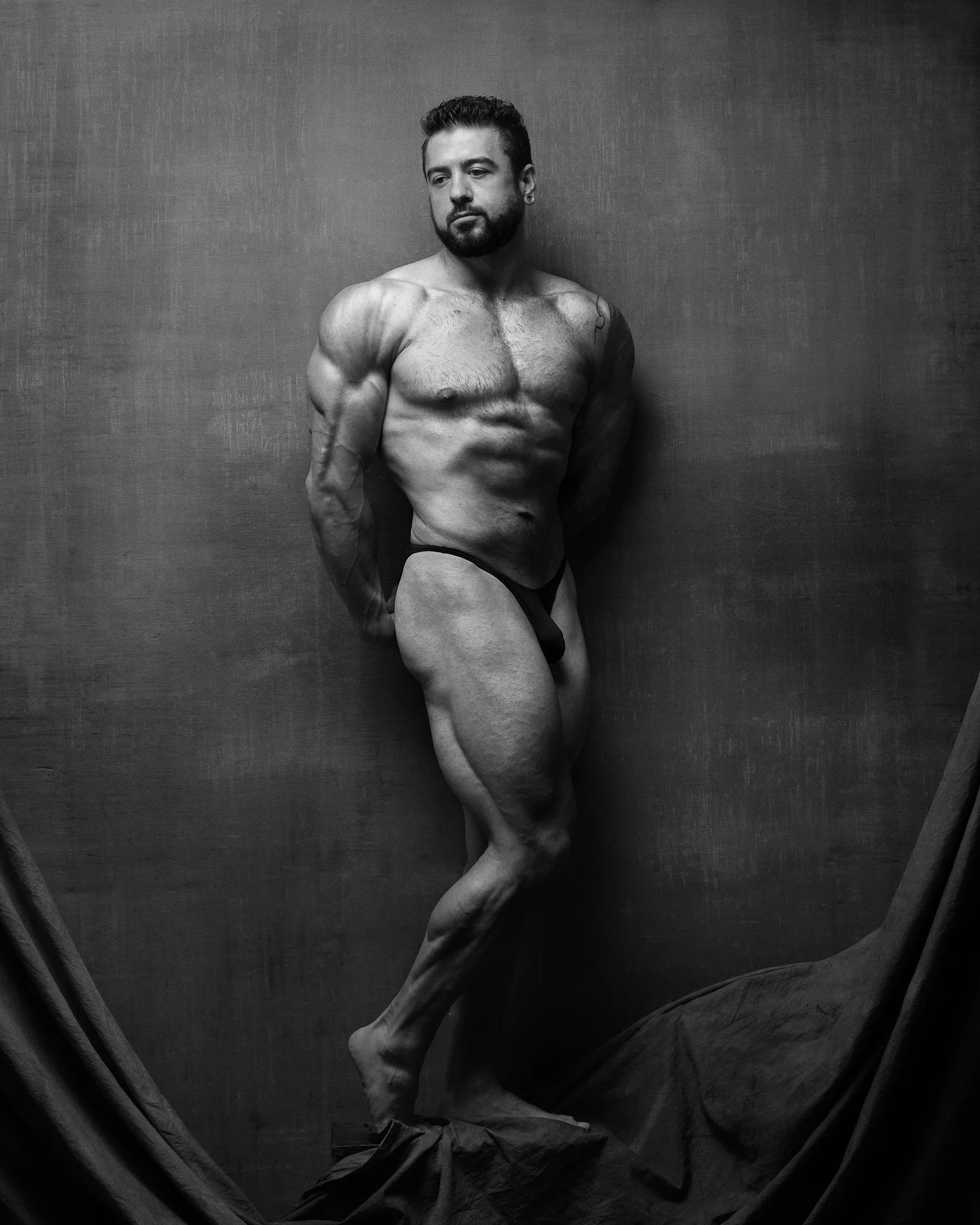 Black and white photo of a muscular man with a beard, partially nude, leaning against a plain wall with draped fabric on the floor.