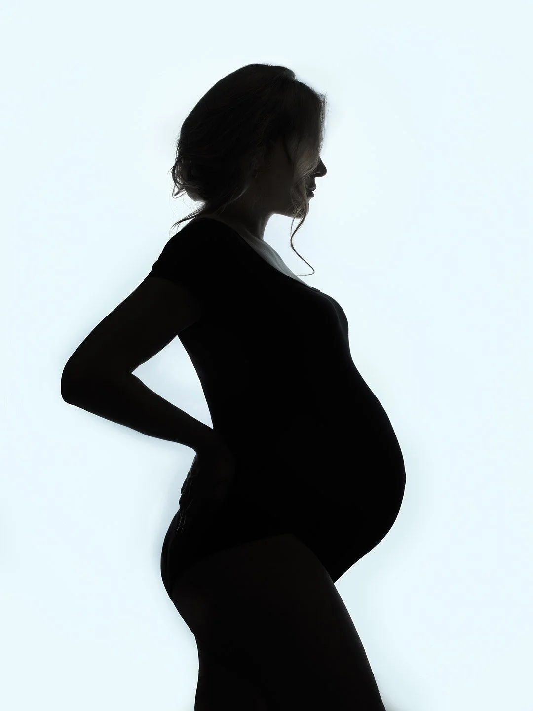 Silhouette of a pregnant woman in profile, standing with her hands on her lower back against a white background.