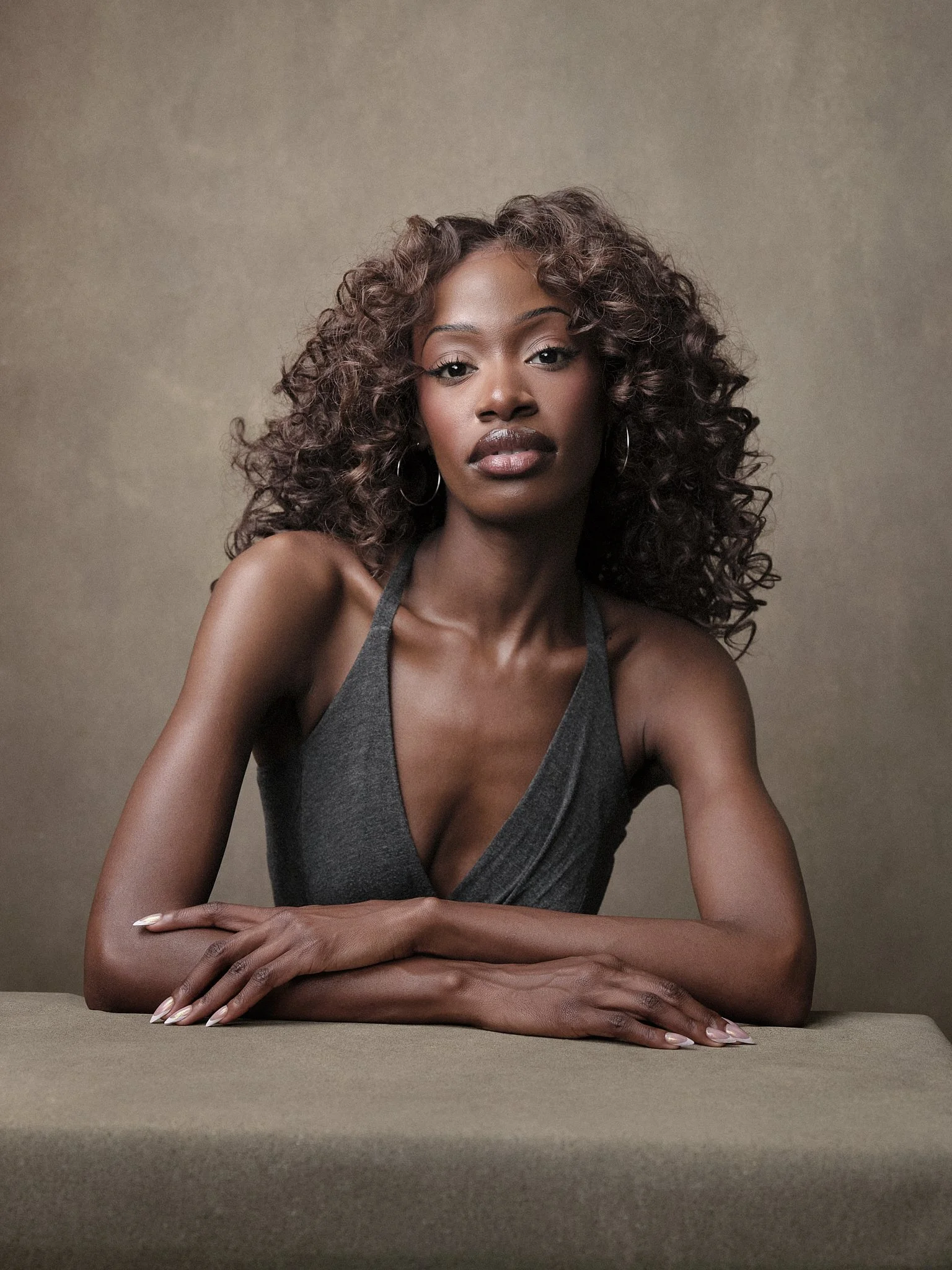 A woman with curly hair sitting at a table against a beige background, wearing a gray sleeveless top and hoop earrings.