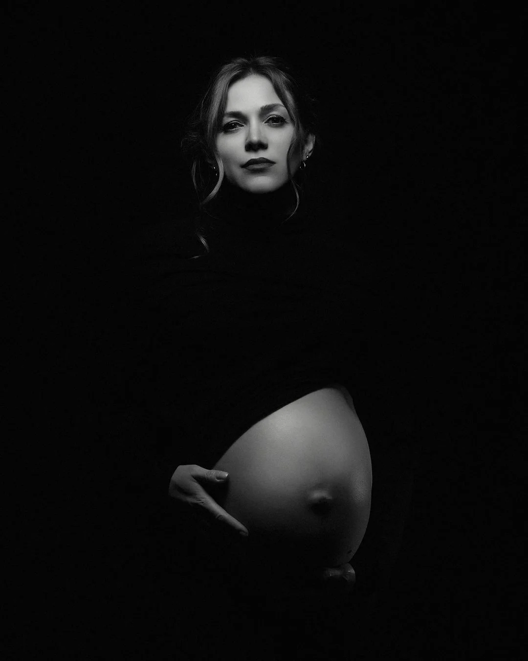 Black and white portrait of a woman with wavy hair and dark lipstick, holding her pregnant belly with one hand, against a dark background.