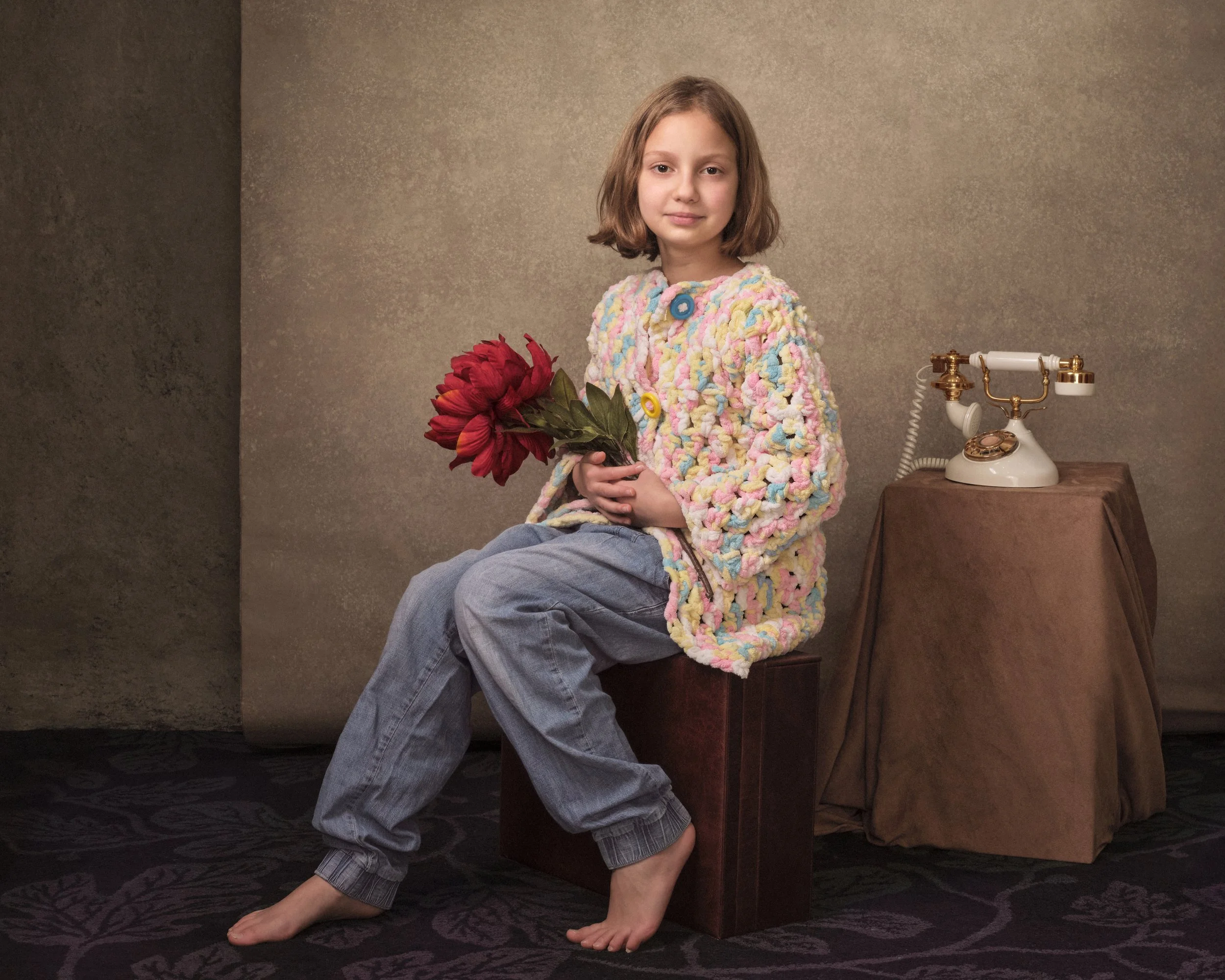 A young girl with shoulder-length light brown hair sitting on a wooden block, holding a large red flower. She is wearing a multicolored, textured coat and gray sweatpants, barefoot. Beside her is a vintage white rotary telephone on a draped brown tab
