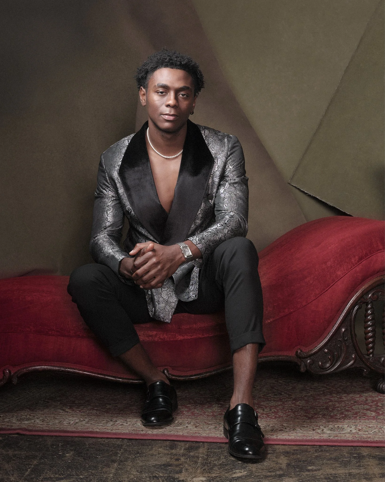 A man dressed in a shiny silver and black blazer, black pants, and black shoes, sitting on a vintage red velvet chaise lounge with a dark, textured background.