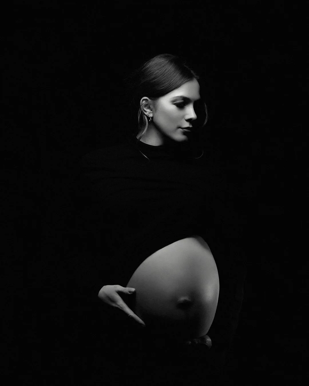 A black-and-white portrait of a pregnant woman with her eyes closed, gently holding her belly, with dark background and soft lighting highlighting her face and belly.