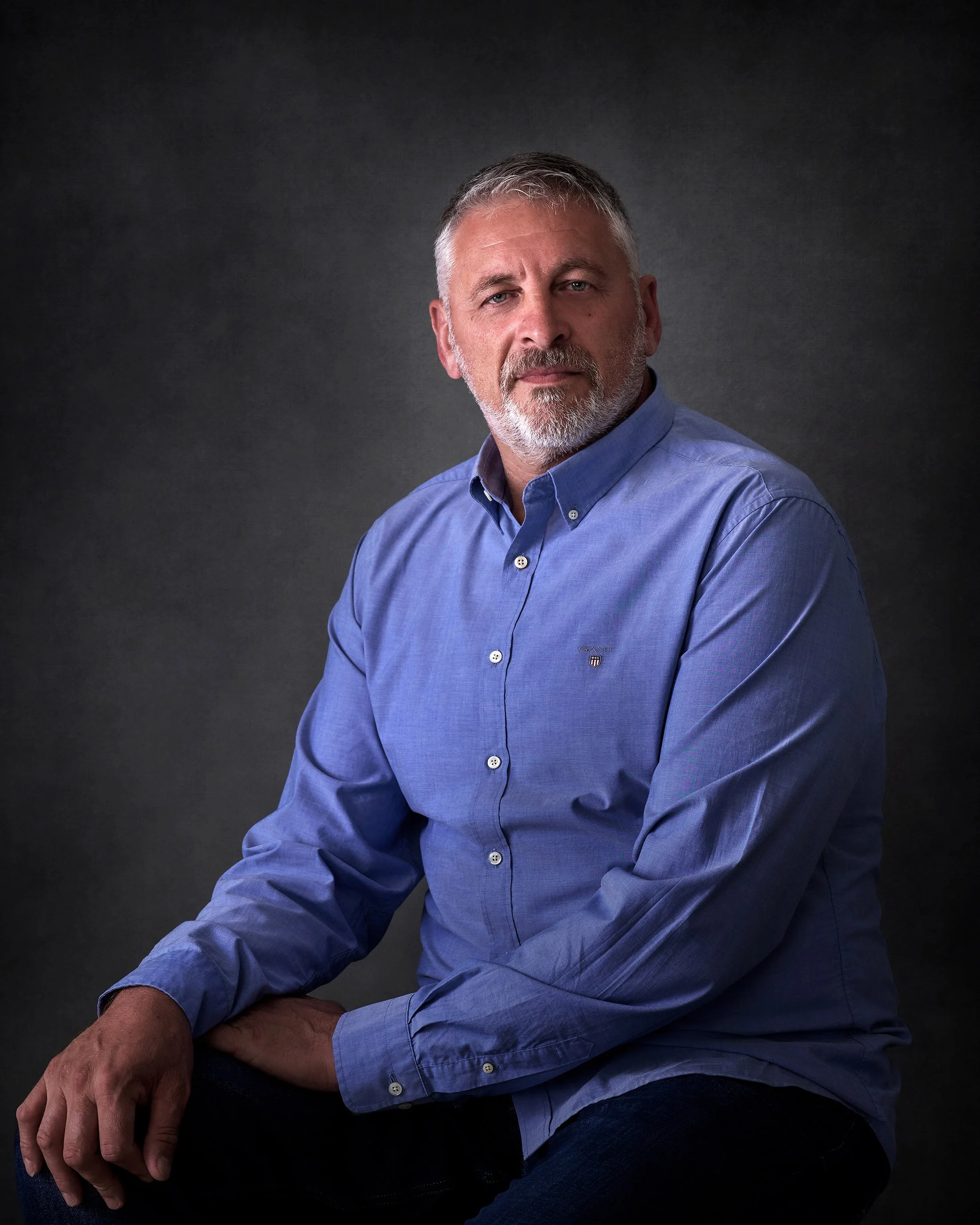 A middle-aged man with gray hair and a beard, wearing a blue button-up shirt, sitting against a dark gray background.
