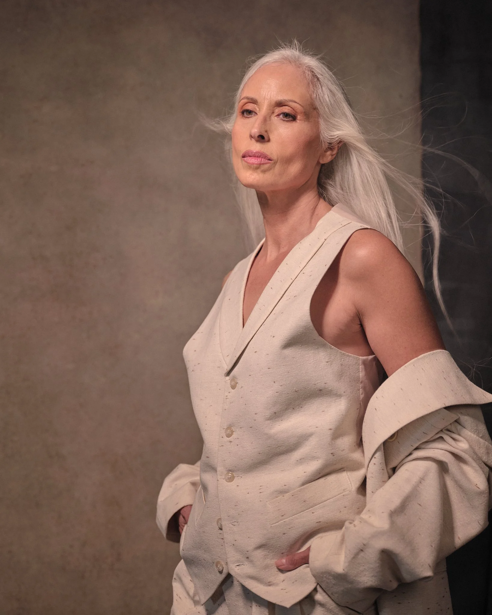 An elderly woman with long gray hair wearing a beige sleeveless vest and matching oversized coat draped over her shoulders, standing against a plain background.