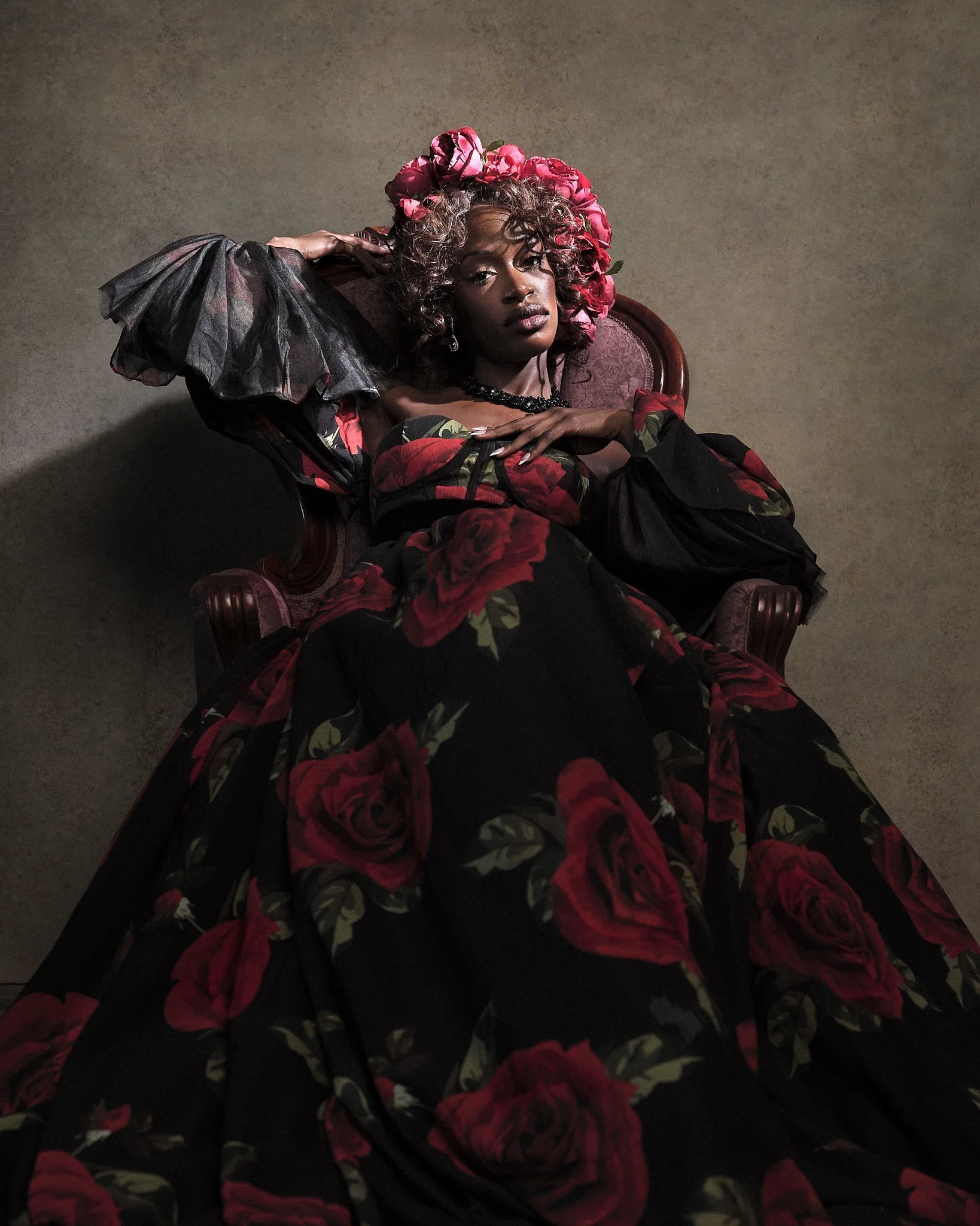 A woman dressed in a long black dress with red roses, lying on a vintage chair, wearing a floral headpiece, with one arm resting on her head and the other on her chest.