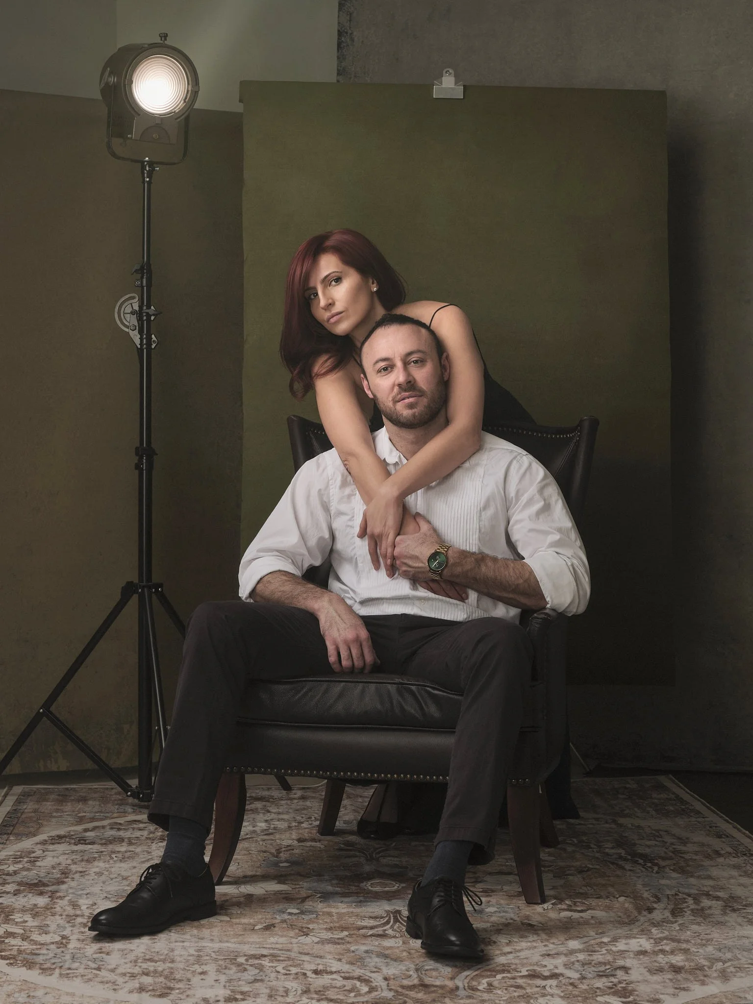 A man with a beard and a woman with red hair posing together in a studio setting. The man is sitting on a black leather armchair, wearing a white shirt and black pants, with one wristwatch visible. The woman is standing behind him, leaning over his shoulder with her arms wrapped around his neck, wearing a black sleeveless top. The background includes a green backdrop and a studio light on a stand to the left.