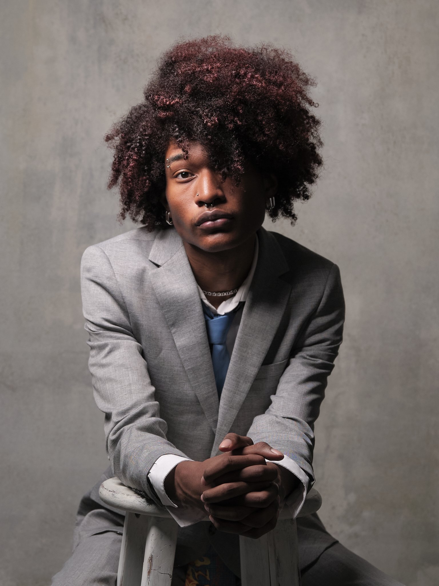 A person with dark skin, curly hair, and wearing a gray suit, sitting on a white stool against a plain background.