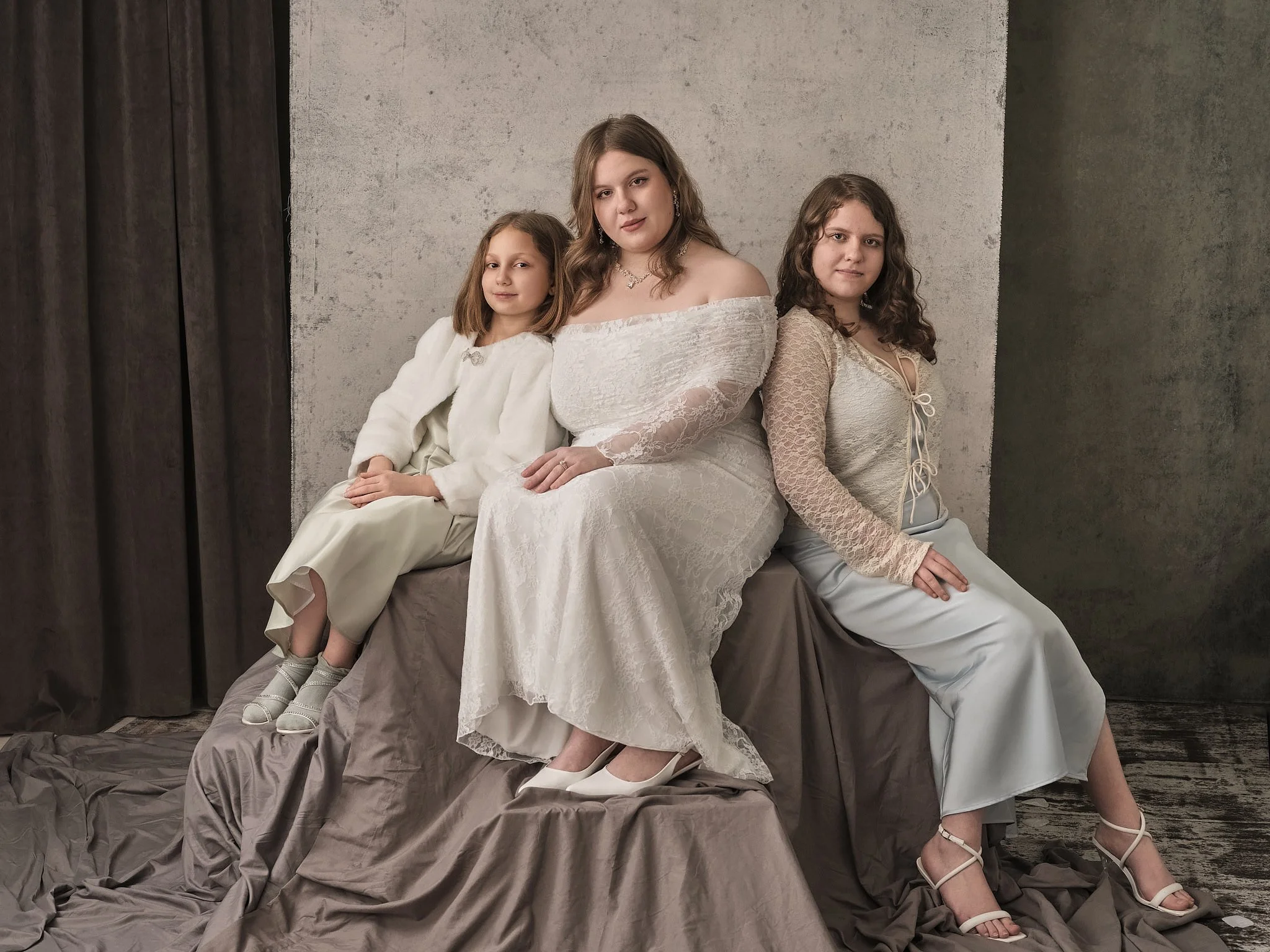 Three women sitting on a draped surface in front of a textured wall, dressed in white attire, with their hands on their laps or resting on the surface.
