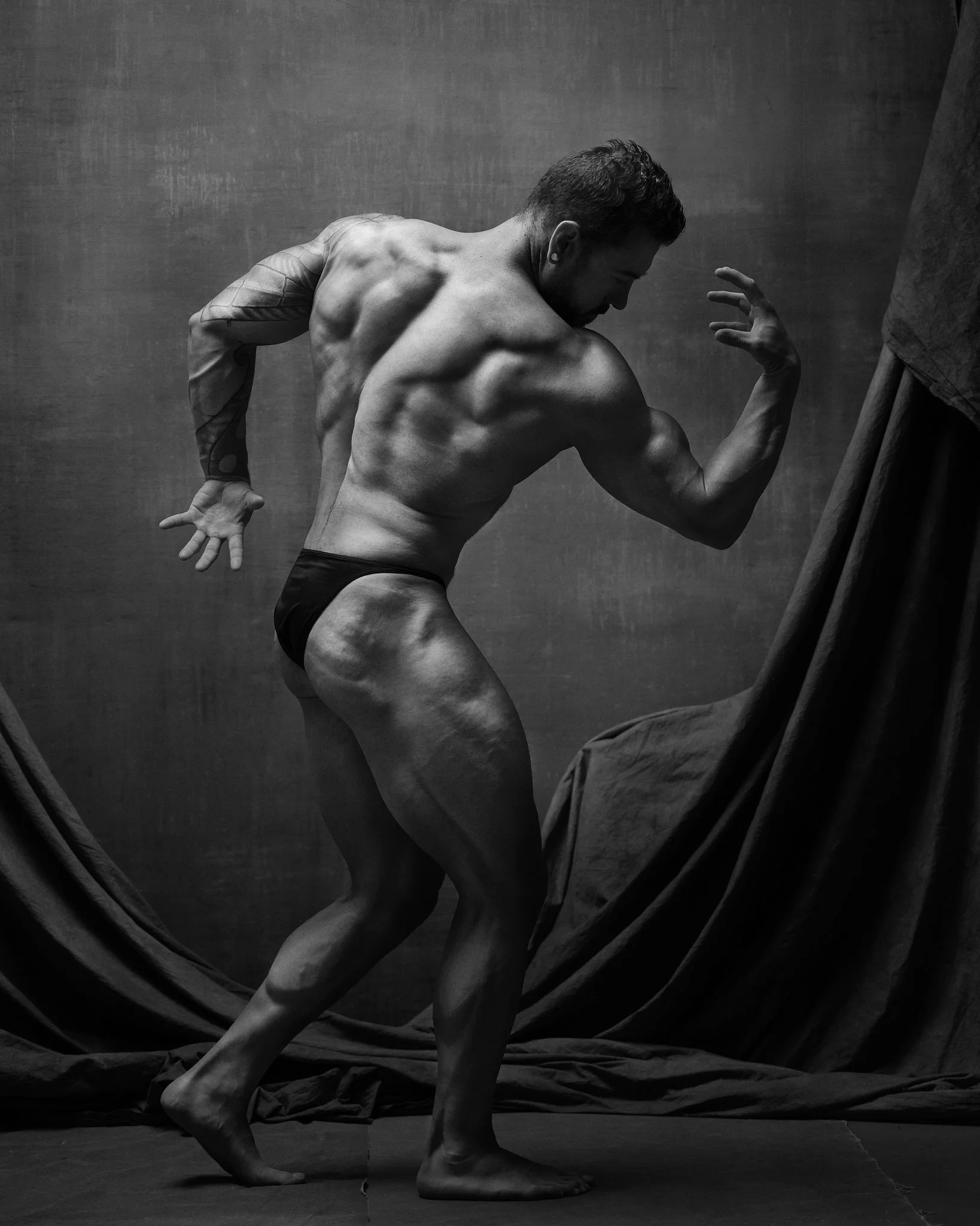 A muscular man in black underwear poses with his back and arm muscles flexed against a dark textured background, partially draped with fabric.