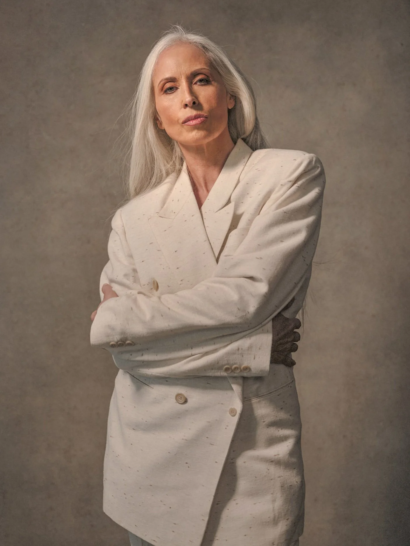 An older woman with long gray hair, dressed in a cream-colored blazer and skirt, standing with arms crossed against a plain background.
