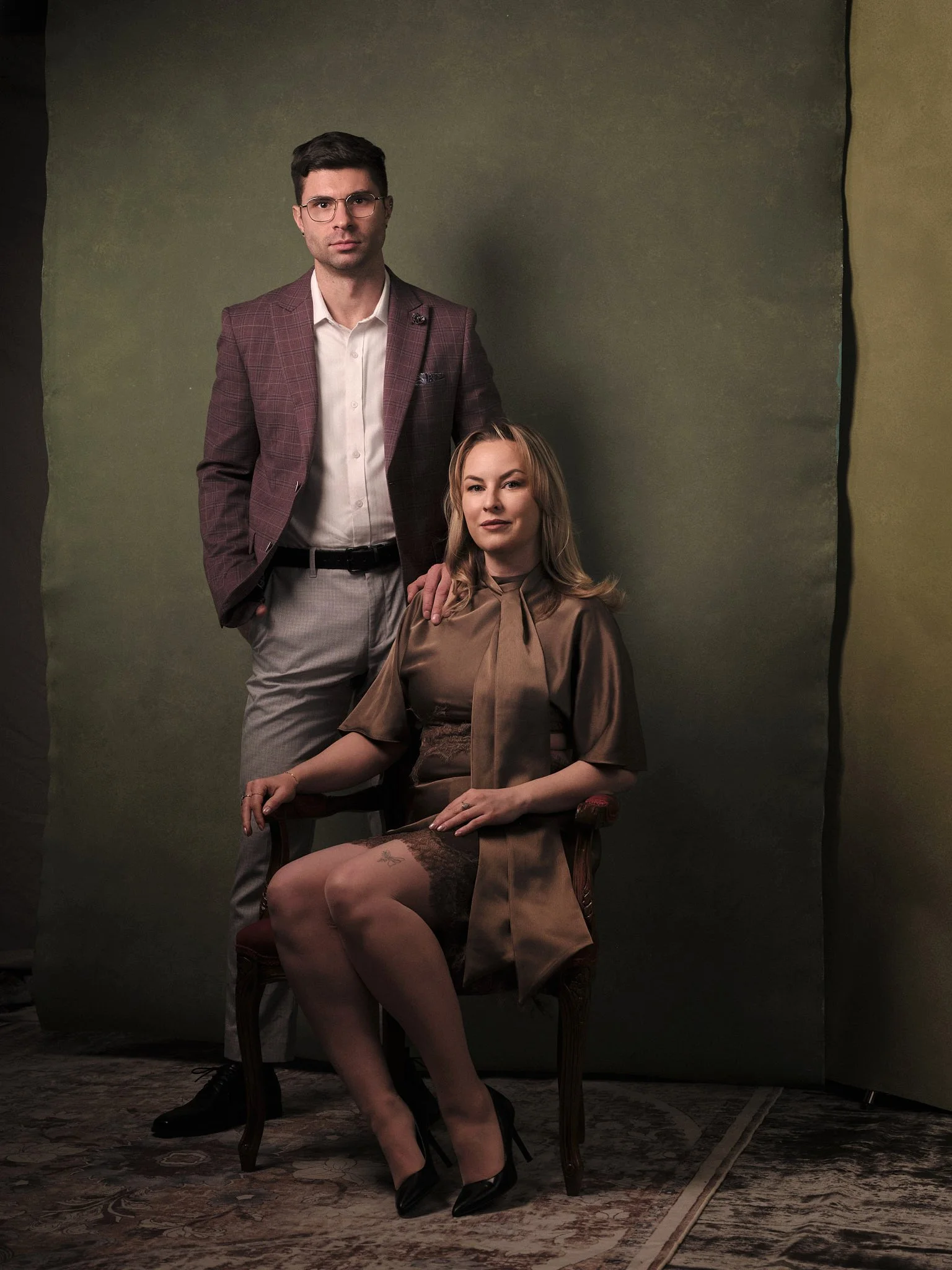 A man and woman pose for a portrait in front of a dark green background. The man is standing, wearing a blazer, white shirt, and gray pants. The woman is seated on a wooden chair, wearing a brown satin dress with lace stockings and black high heels, with her hair styled in loose waves.