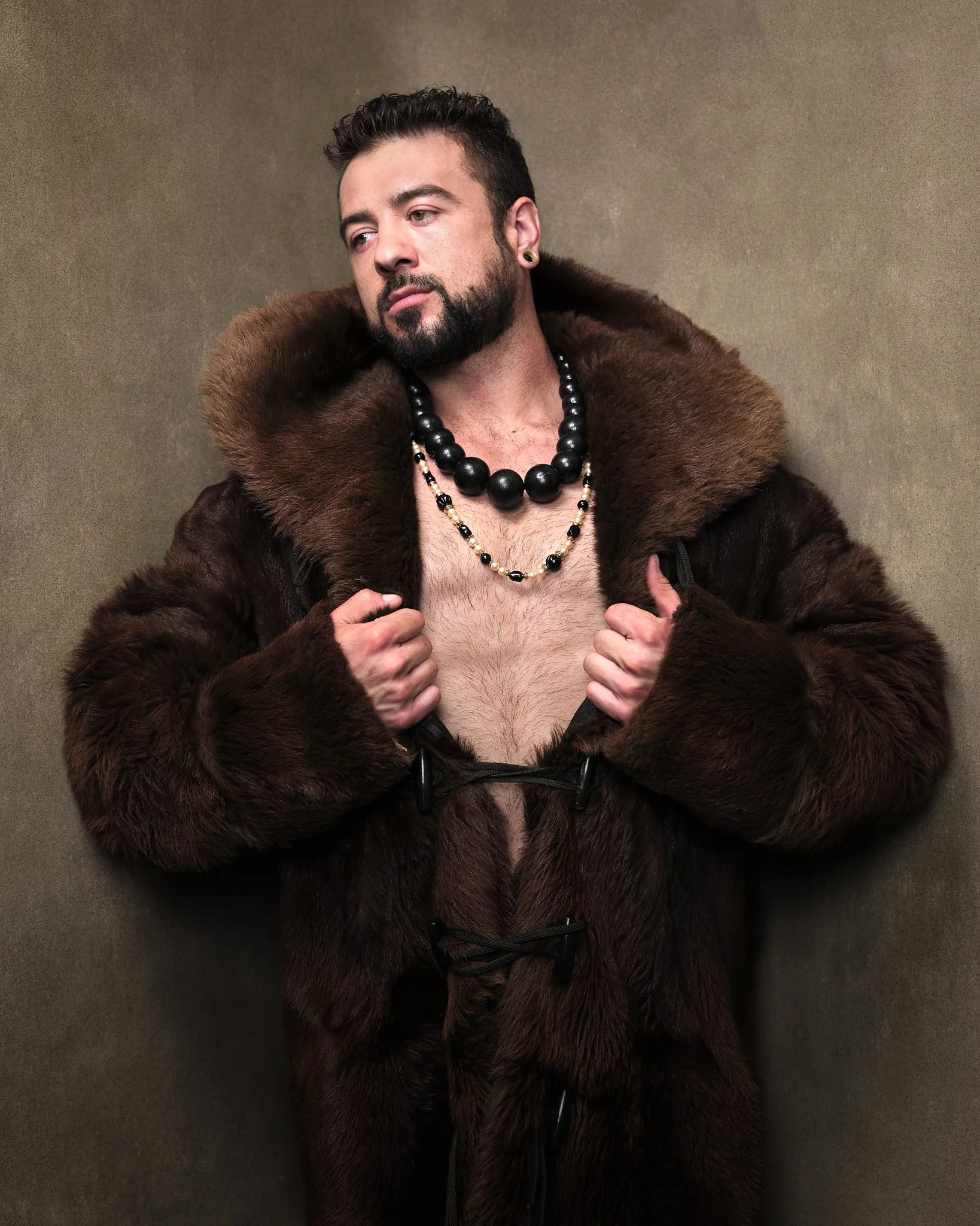 A man with a beard and styled hair wearing a large fur coat, unbuttoned to reveal his chest, and accessorized with black and gold beaded necklaces, earrings, and a ring.