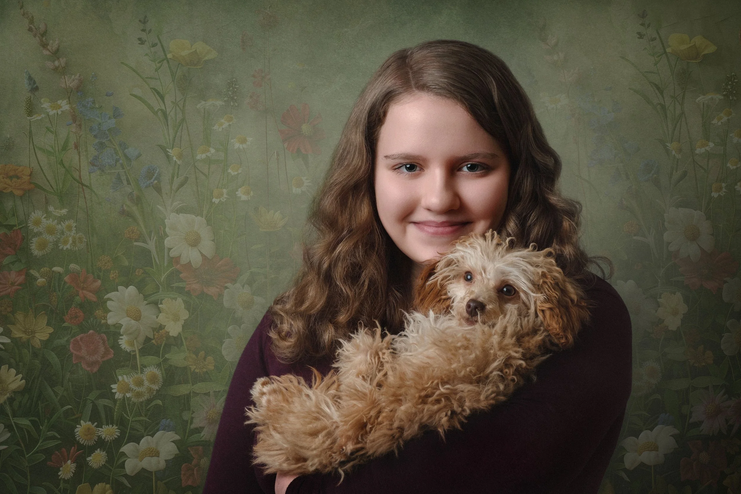 A girl with long curly brown hair holding a small curly-haired puppy in front of a floral background.