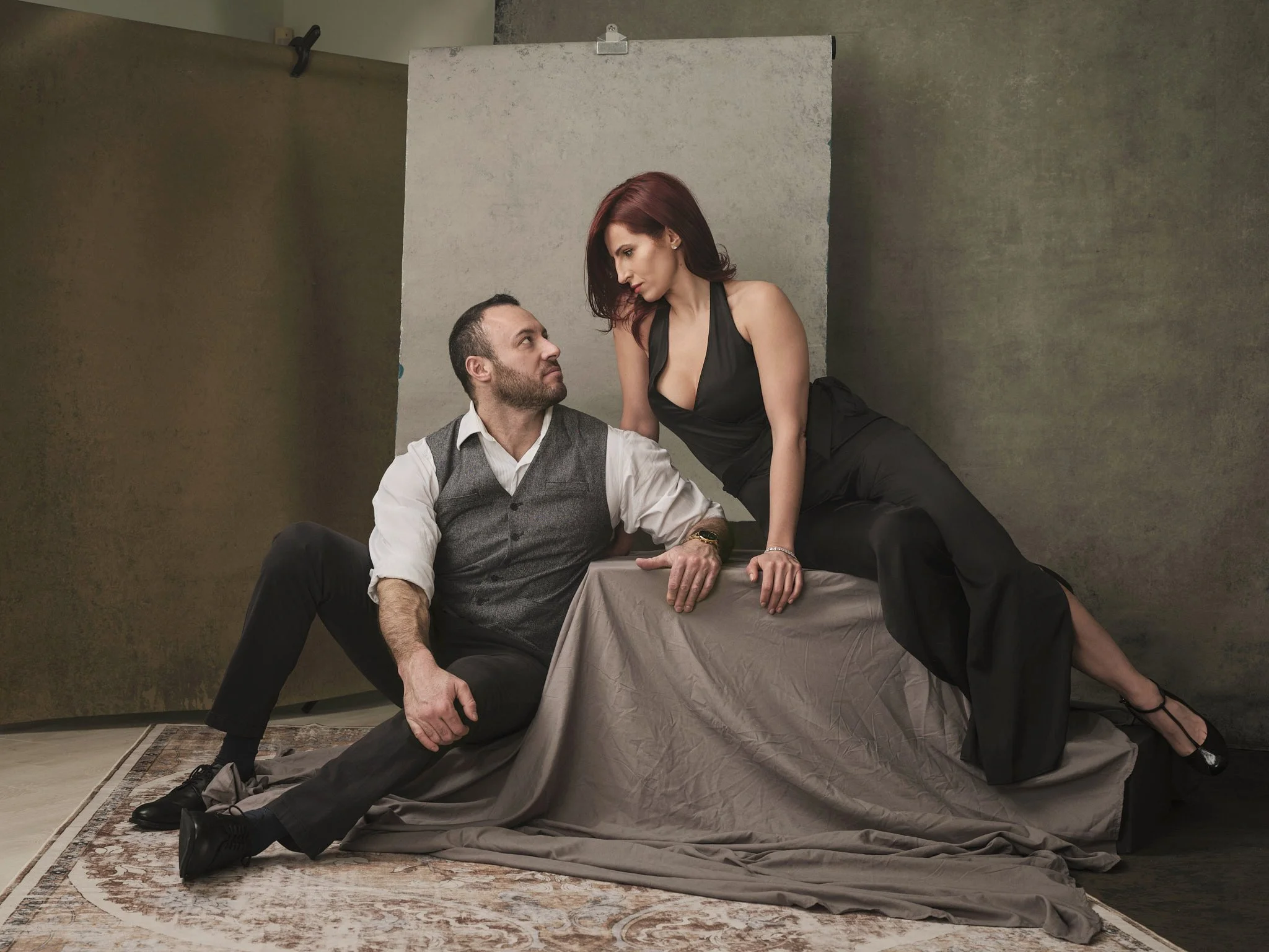 A man and a woman posing together in a studio setting with a plain background. The man is seated on the floor, leaning on a draped cloth, looking up at the woman. The woman is standing with one leg on a surface, leaning towards the man, looking down 