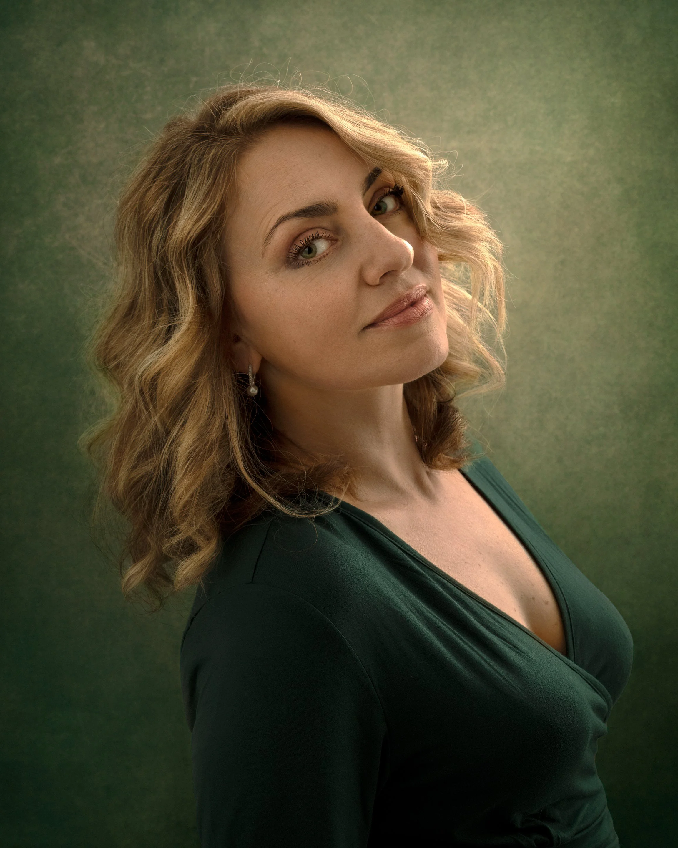 A woman with wavy blonde hair and green eyes, wearing a dark green top, looking at the camera with a slight smile, against a textured green background.