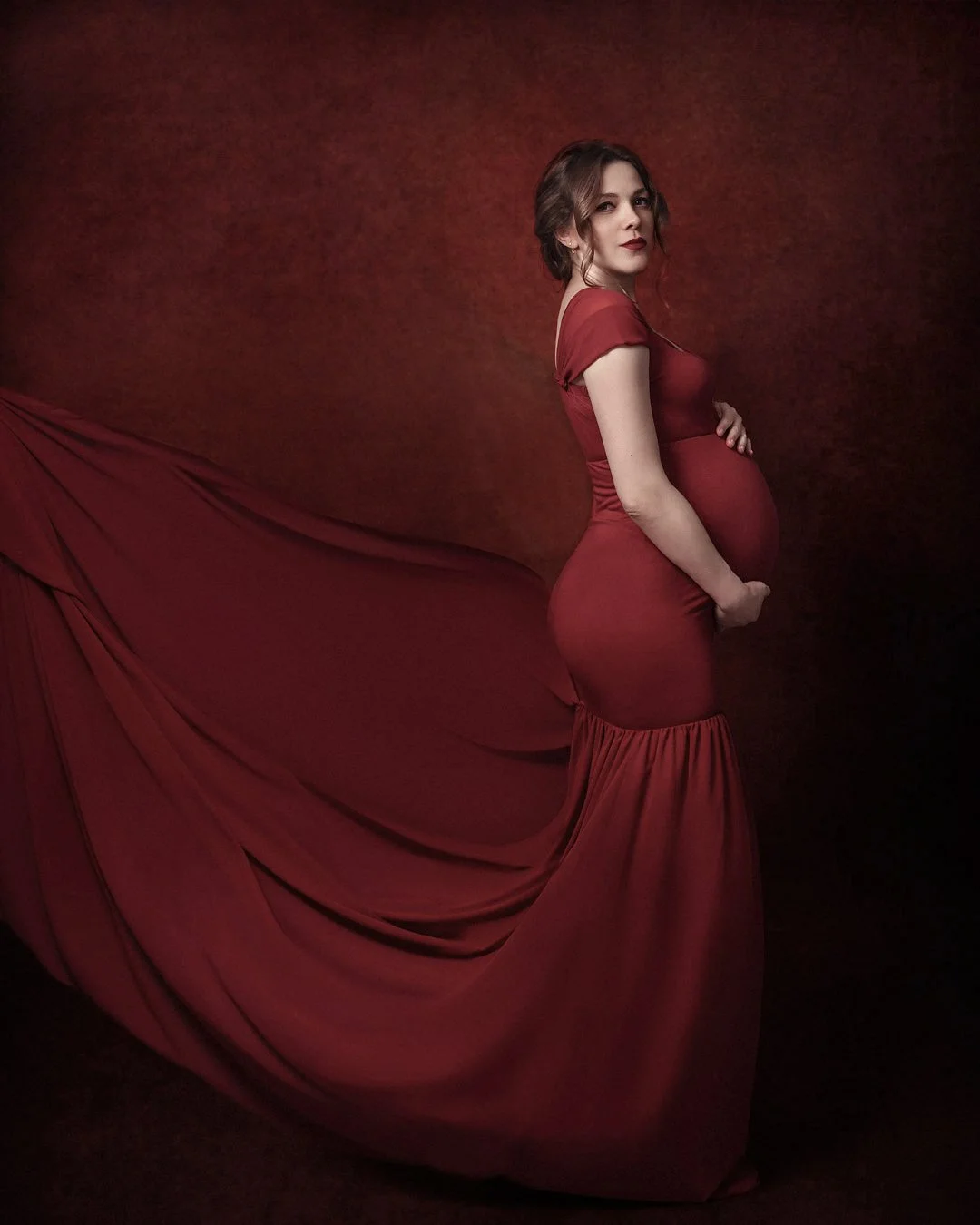 Pregnant woman in a red dress with a flowing train, standing against a matching red background.