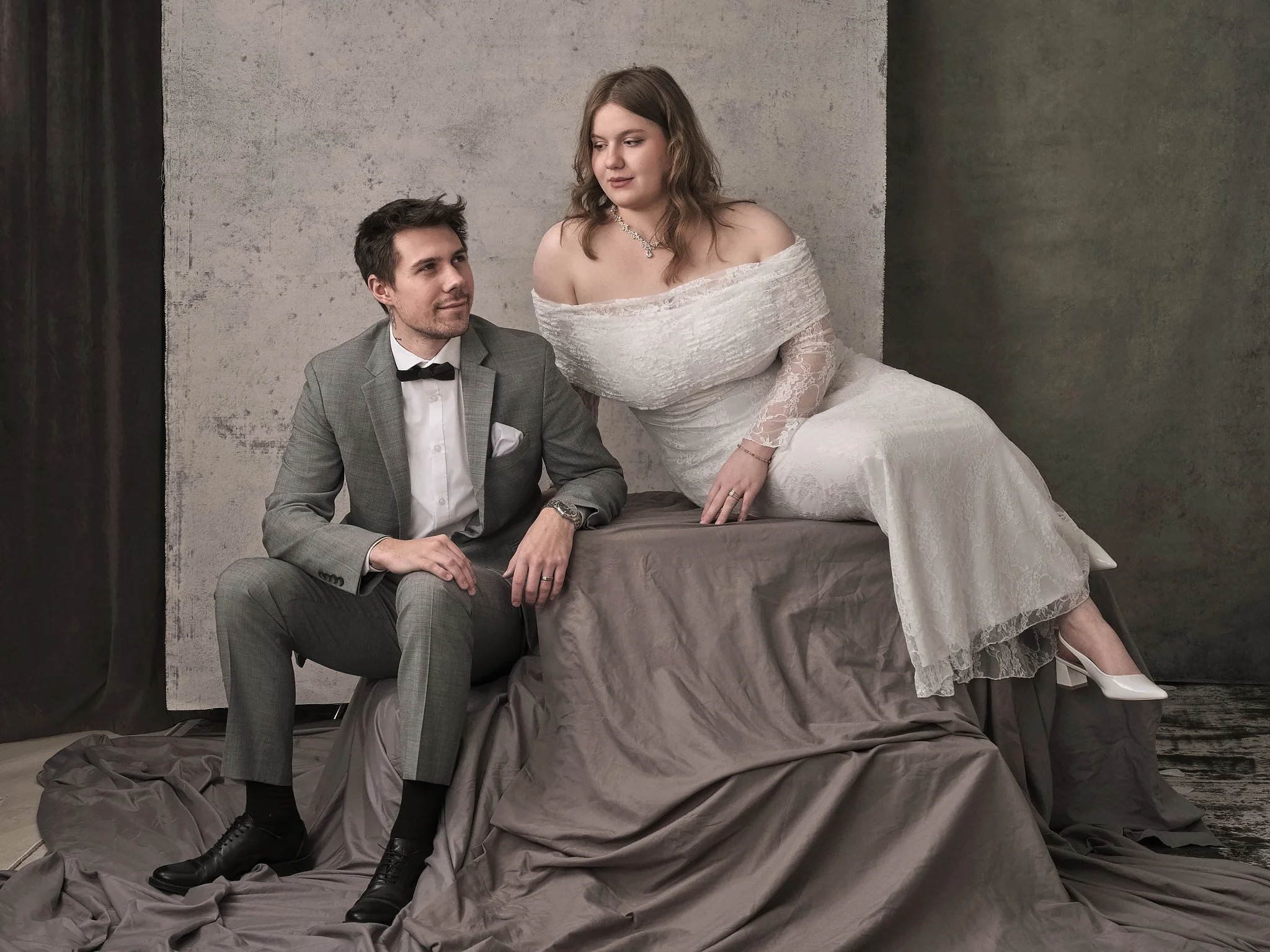 A man and woman in wedding attire pose for a photo in front of a plain background. The woman is sitting on a draped surface, leaning toward the man who is sitting beside her. The woman is wearing a white lace wedding gown and white heels, while the m