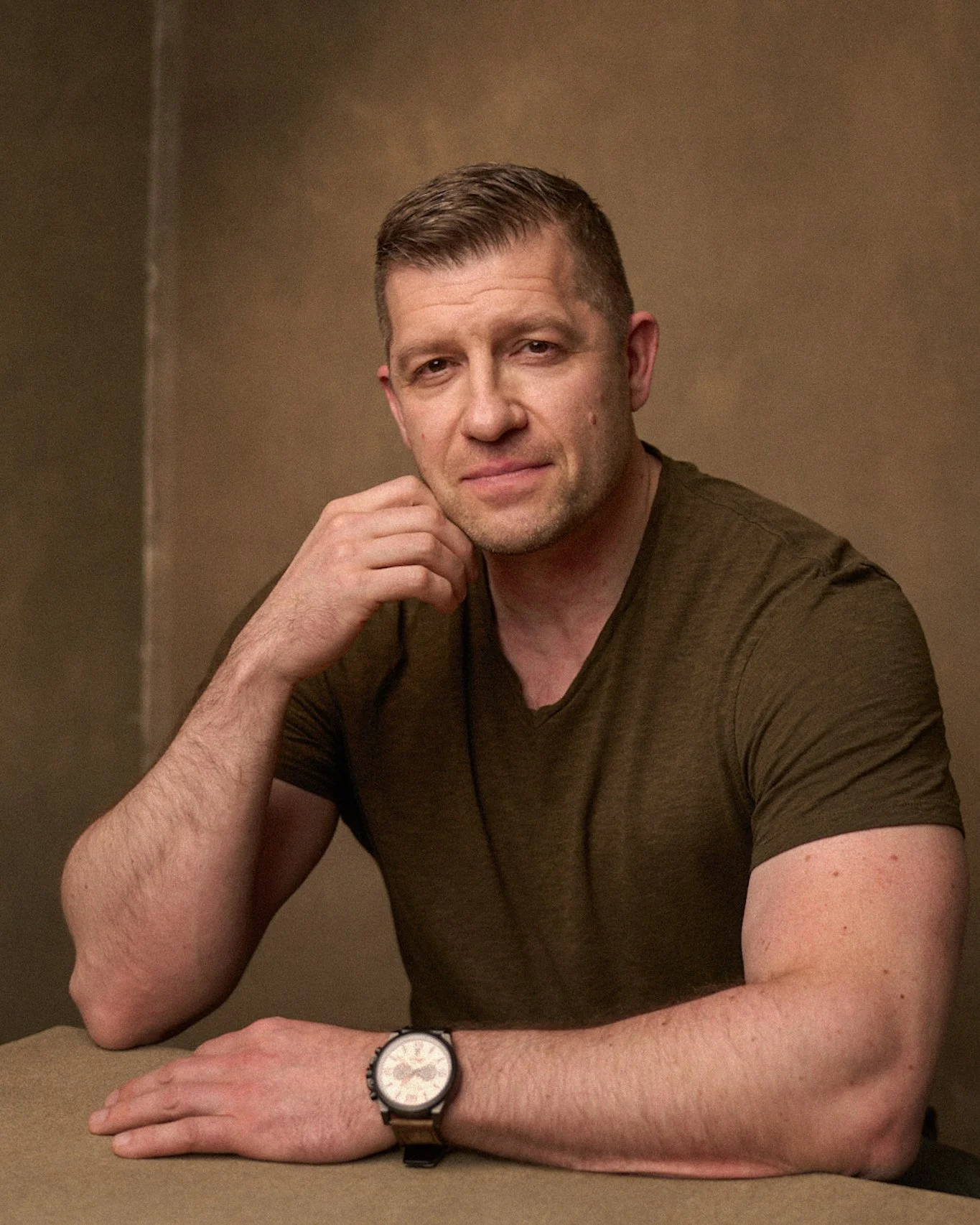A man in a dark green T-shirt, with short brown hair, sitting at a table with his left arm resting on the surface. He is wearing a watch and has a slight smile, with a plain beige background.