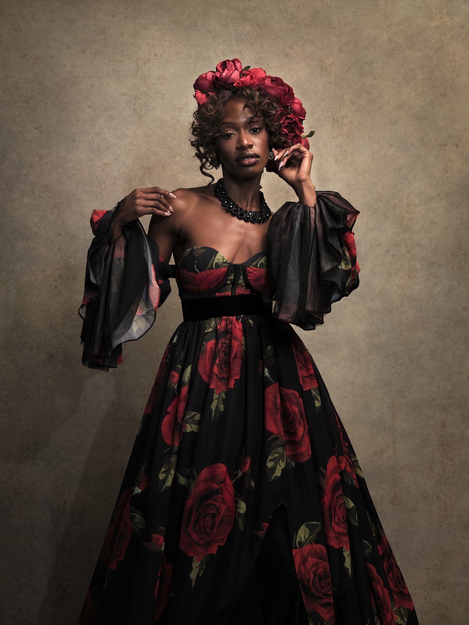 A woman wearing a black and red floral dress with matching floral headpiece, black choker, and earrings, standing against a textured beige background.