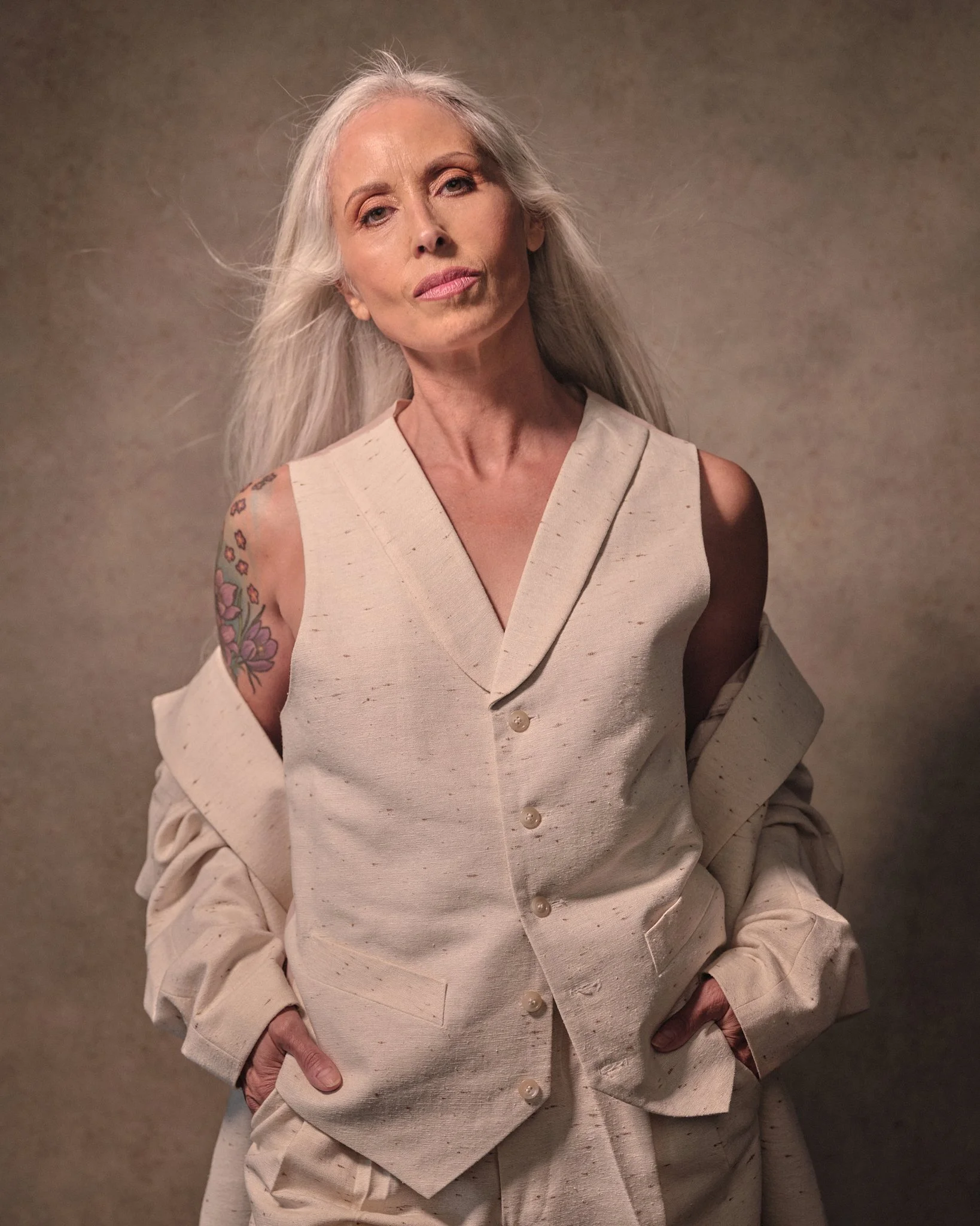 An elderly woman with long, gray hair stands against a plain background, wearing a cream-colored, sleeveless outfit with a matching jacket off her shoulders. She has a colorful tattoo on her right upper arm and looks confidently into the camera.