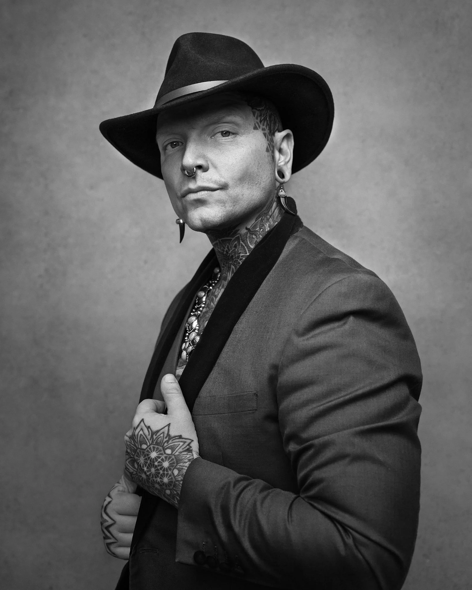 A man with tattoos, wearing a hat and a blazer, looking at the camera against a plain background.