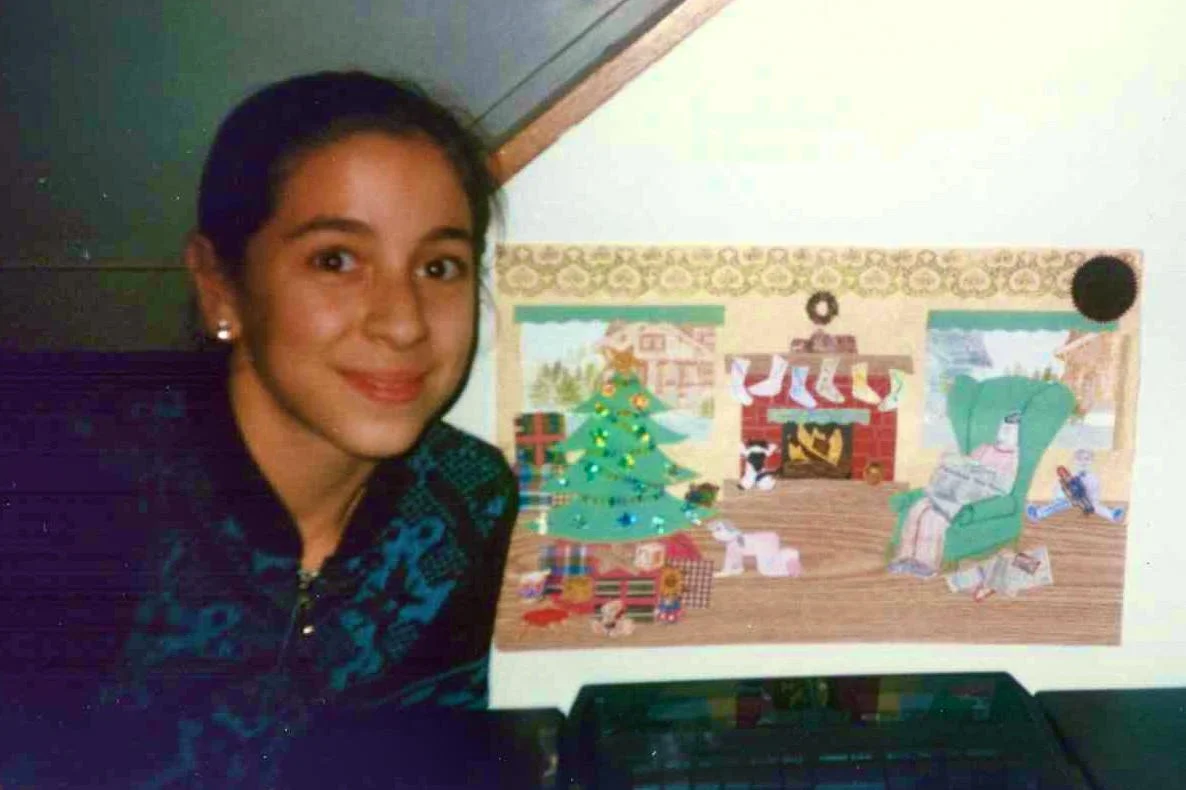 Me smiling as a kid sitting next to a Christmas illustration with a decorated Christmas tree, fireplace with stockings, and wrapped gifts that I made. It won a contest with a local newspaper.