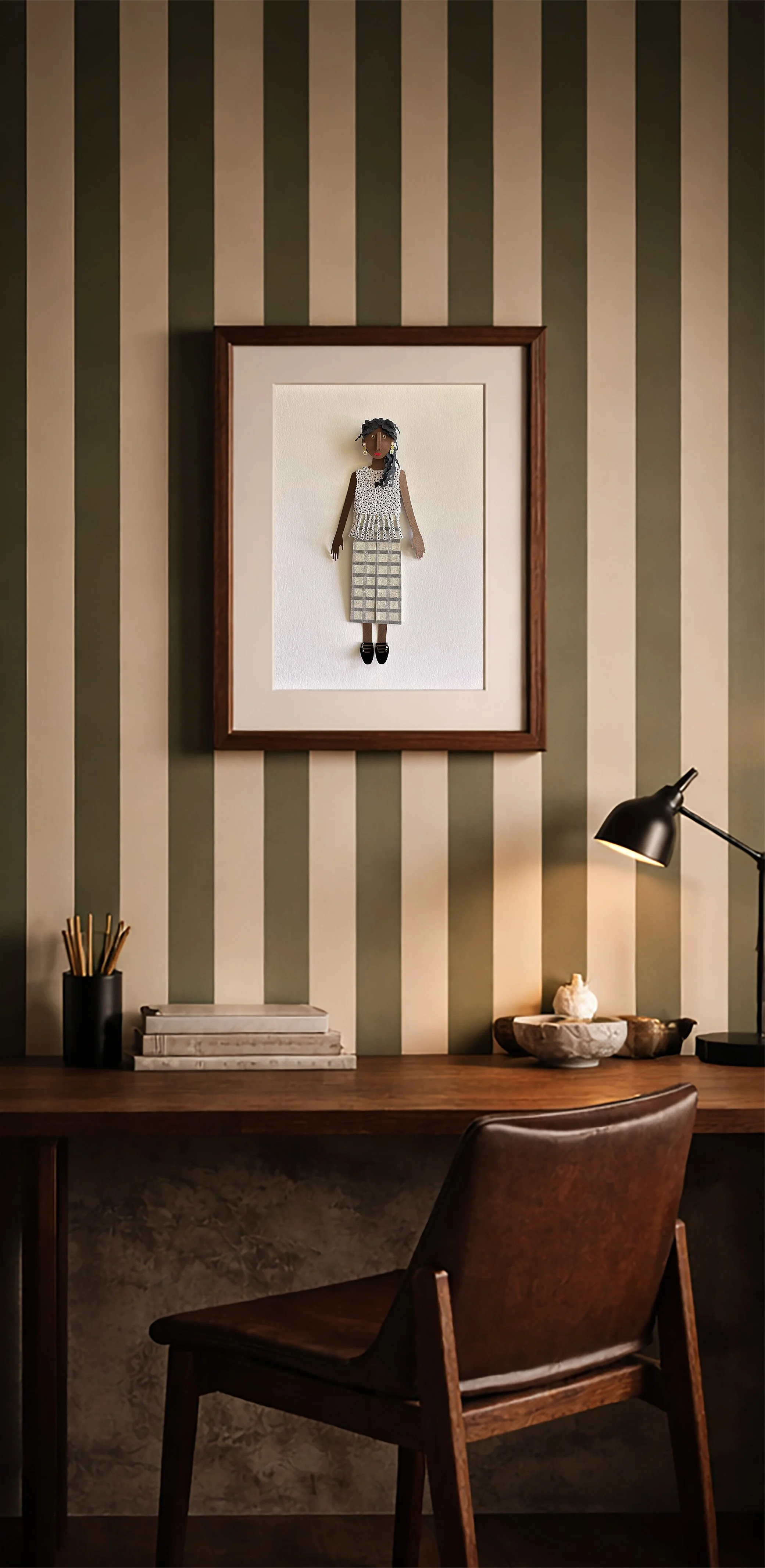 A home office setup featuring a wooden desk and chair in front of a wall with green and beige vertical stripes. Above the desk, a framed Brittani Rose original paper girl art piece is displayed.