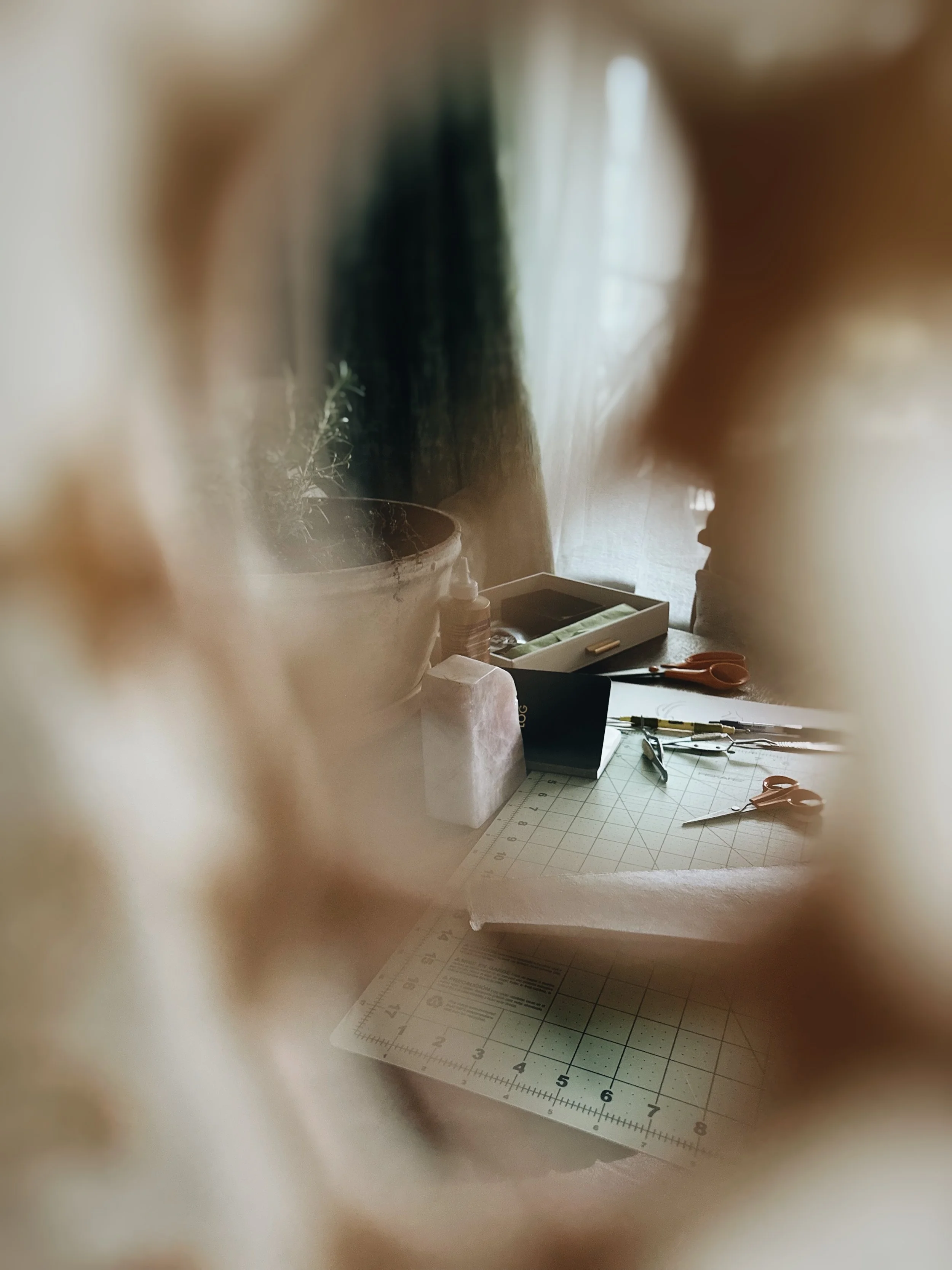 A view through a small, irregularly shaped opening shows a workspace with scissors, a cutting mat, and various crafting tools, with a window and curtain in the background.