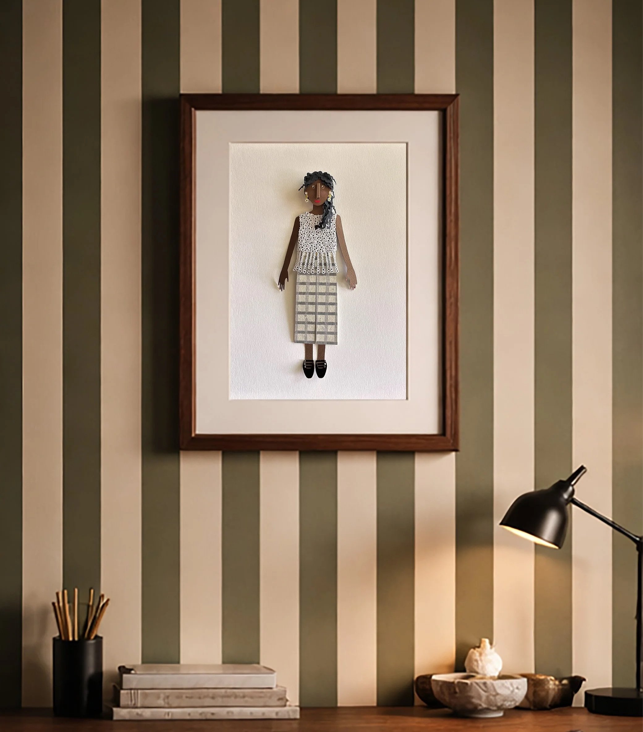 Decorative framed textile art of a woman in a plaid pants and white floral top hanging on a wall with green and beige striped wallpaper, and a desk with books, a pen holder, and decorative objects, a black desk lamp, and a white vase.
