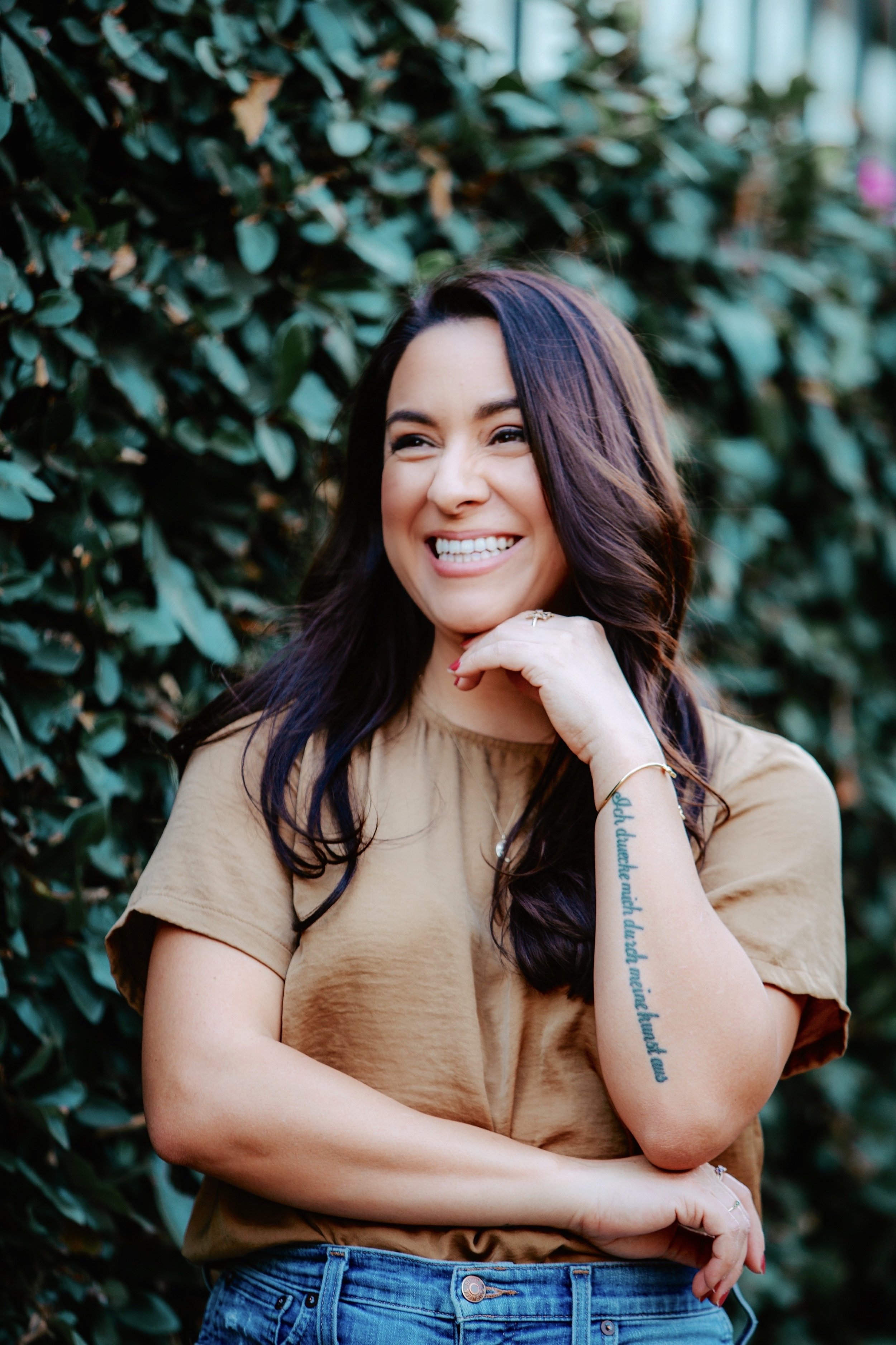A woman with long dark hair smiling, standing outdoors in front of a bush with green leaves, wearing a tan top and blue jeans.